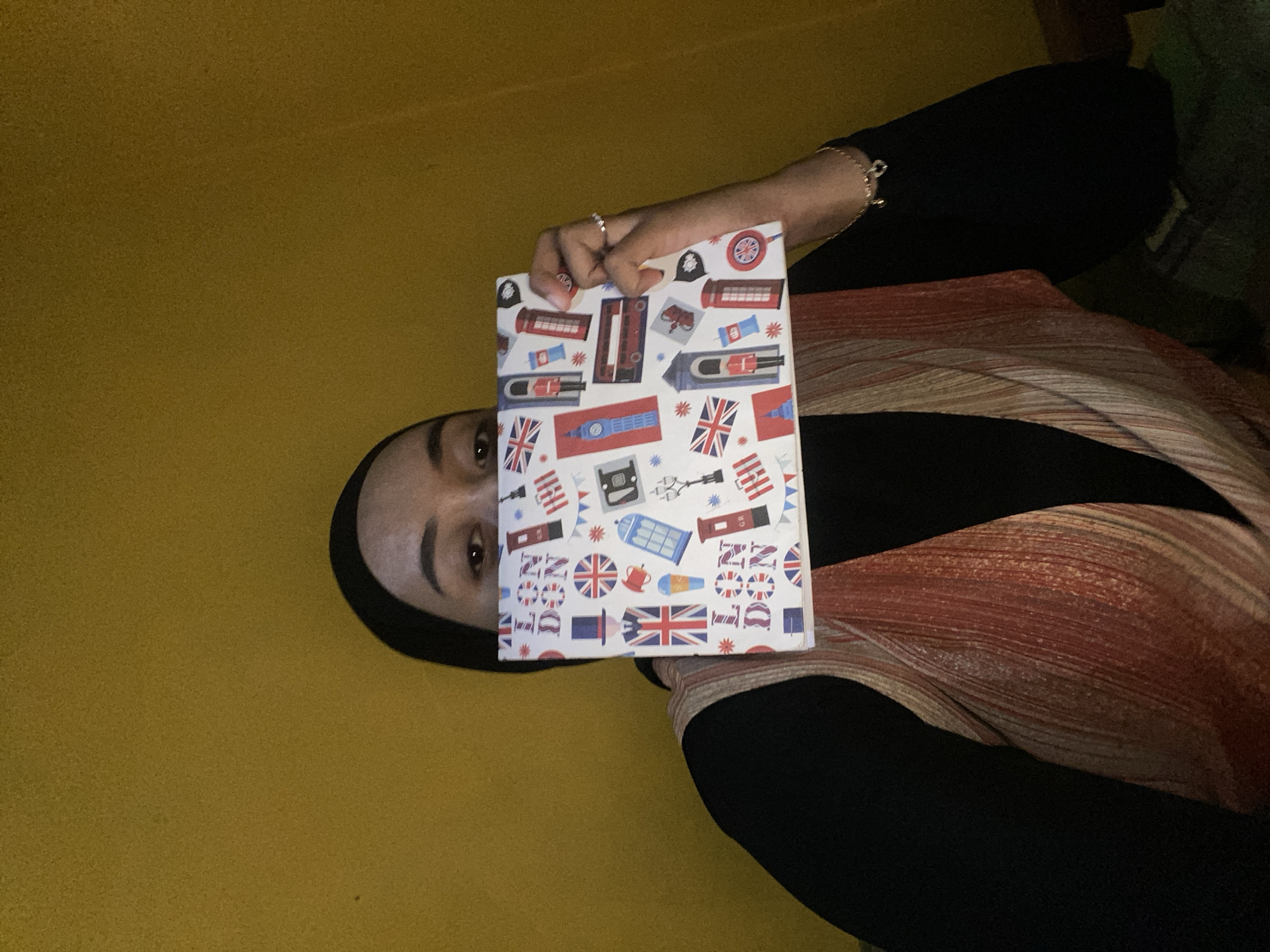 A person wearing a black hijab and a striped vest over a black top holds up a rectangular object with a London-themed pattern, obscuring their face. The object is decorated with images of Union Jacks, red phone booths, Big Ben, a double-decker bus, and other London landmarks. The background is a textured yellow wall. The person is wearing a ring on their right hand and a bracelet with charms on their left wrist. The scene appears to be indoors, possibly in a residential setting. The image does not provide enough information to determine the location in Kecamatan Pati, Indonesia.