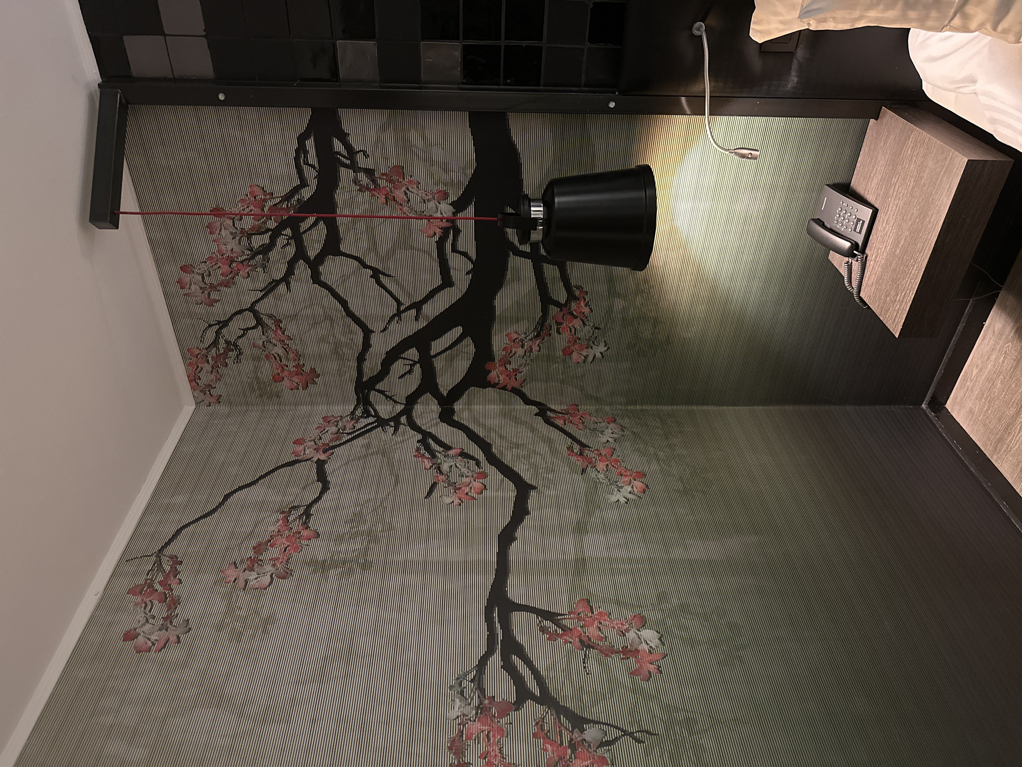 The image depicts a hotel room interior. A black wall-mounted lamp is visible, with its cord running across the wall. The wall features a wallpaper with a pattern resembling a black tree with pink blossoms against a striped background. A telephone is placed on a wooden bedside table. There is a small flexible reading light fixed to the same table. The location is likely in Genk, Belgium, but that cannot be verified from the image itself. No people are visible.