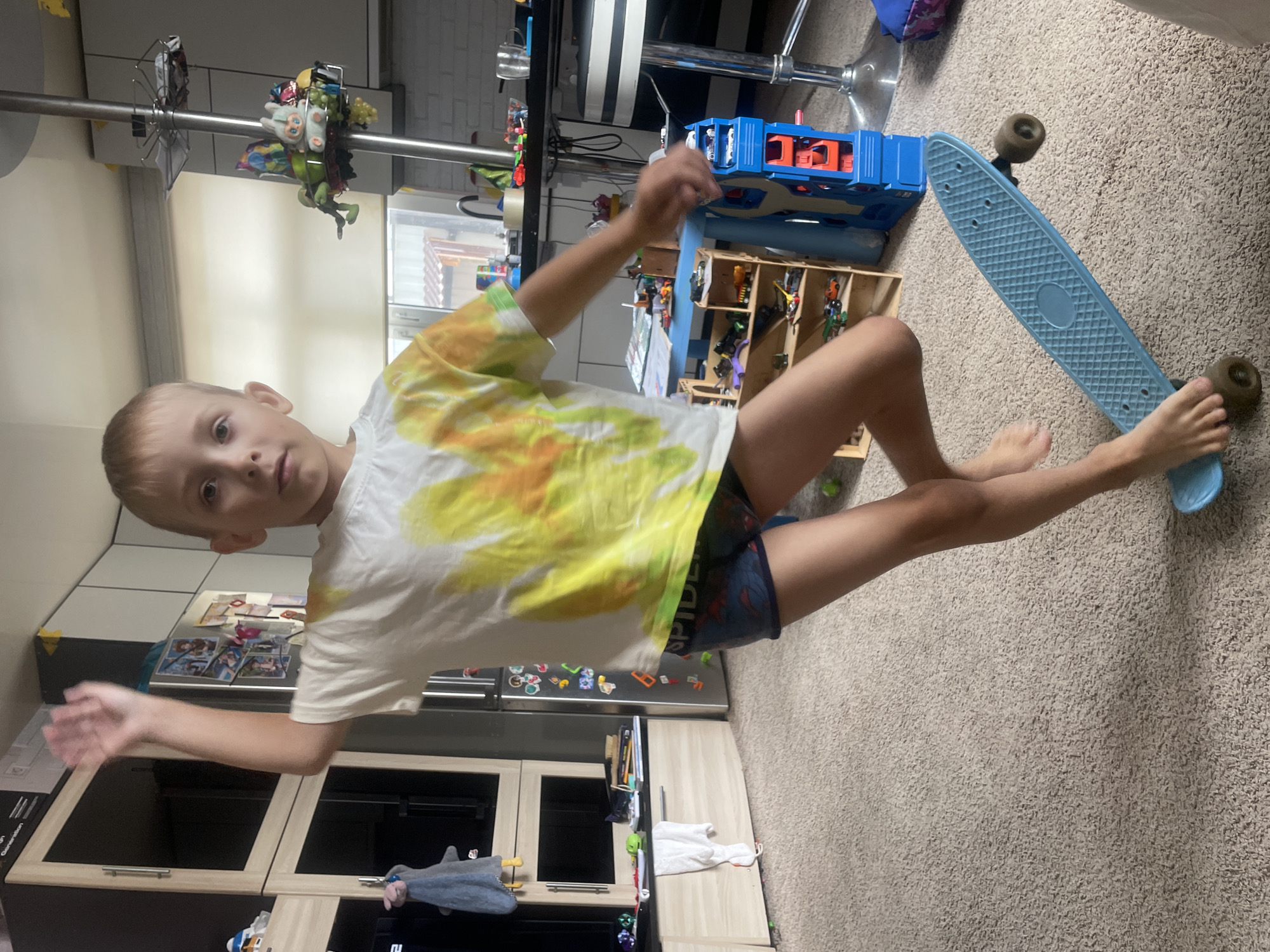A young boy with blonde hair and light skin stands on a blue penny board in a room. He is wearing a tie-dye t-shirt and blue patterned shorts. The room appears to be a home, with a kitchen area in the background, including cabinets and a refrigerator. There are toys scattered around, including a toy car ramp and shelves filled with miniature vehicles. The floor is carpeted, and a large cabinet is visible to the left of the boy. The lighting suggests it is daytime, possibly with natural light coming from a window not visible in the frame. There is no indication of the specific location being Baranovka, Russia.
