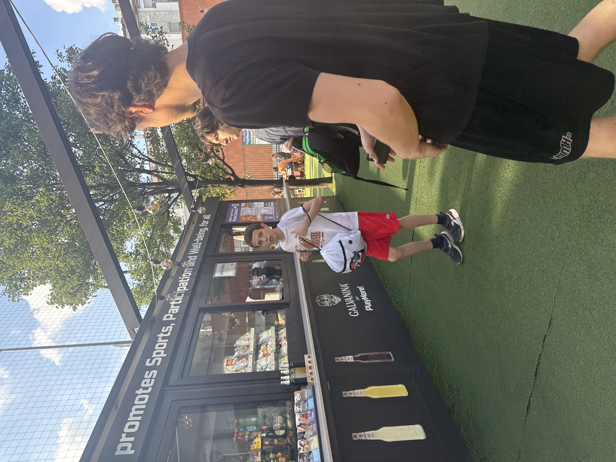 This image depicts an outdoor recreational area in Milan, Italy, specifically identified by the "PlayMore!" and "Galvanina" branding on a concession stand. The scene is illuminated by natural daylight. In the foreground, a young male, estimated to be in his late teens or early twenties, is positioned on the right, viewed from behind and slightly to his left. He has dark, wavy hair and is wearing a black t-shirt and black shorts with a white "NTH 23" logo on the left leg. He holds a dark-colored smartphone in his left hand. Immediately to his left and slightly behind, another young male, also with dark hair, is visible from the chest up, wearing a grey t-shirt and carrying a black and green backpack over his left shoulder. His head is turned to the left. In the middle ground, to the left of the two young males, a boy, estimated to be 8-12 years old, faces slightly towards the right of the frame, looking towards the camera. He has short, dark hair and is wearing a white t-shirt with a "BASKET ADRIATIC" graphic, red shorts, dark sneakers with white soles, and dark socks. He carries a white drawstring bag with a red and black logo over his left shoulder. To the left, a black concession stand or kiosk extends horizontally across the frame. Its upper section features large white text that reads: "promotes Sports, Participation and Well-being, for all". Below this, suspended string lights with exposed bulbs are visible. The kiosk has multiple glass windows displaying products: the left window shows bottled beverages and snacks, the middle window displays bags of chips (including "San Carlo Patachips") and other packaged snacks, and the right window shows more packaged items and a sign labeled "INFO CORSI". Below the windows, a counter displays brochures and a small wooden stand with bottles. The lower section of the kiosk features large graphic representations of three bottles (white, yellow, dark brown) and the white "GALVANINA per PlayMore!" logo with a skull-like emblem. The ground is covered with artificial green turf, featuring a distinct dark horizontal seam in the lower left-middle. In the background, beyond the kiosk, green trees with dense foliage are prominent, and a light-colored building and a red wall are visible. The upper left portion of the image shows a blue sky with scattered white clouds, partially obscured by a wire mesh fence and a tall