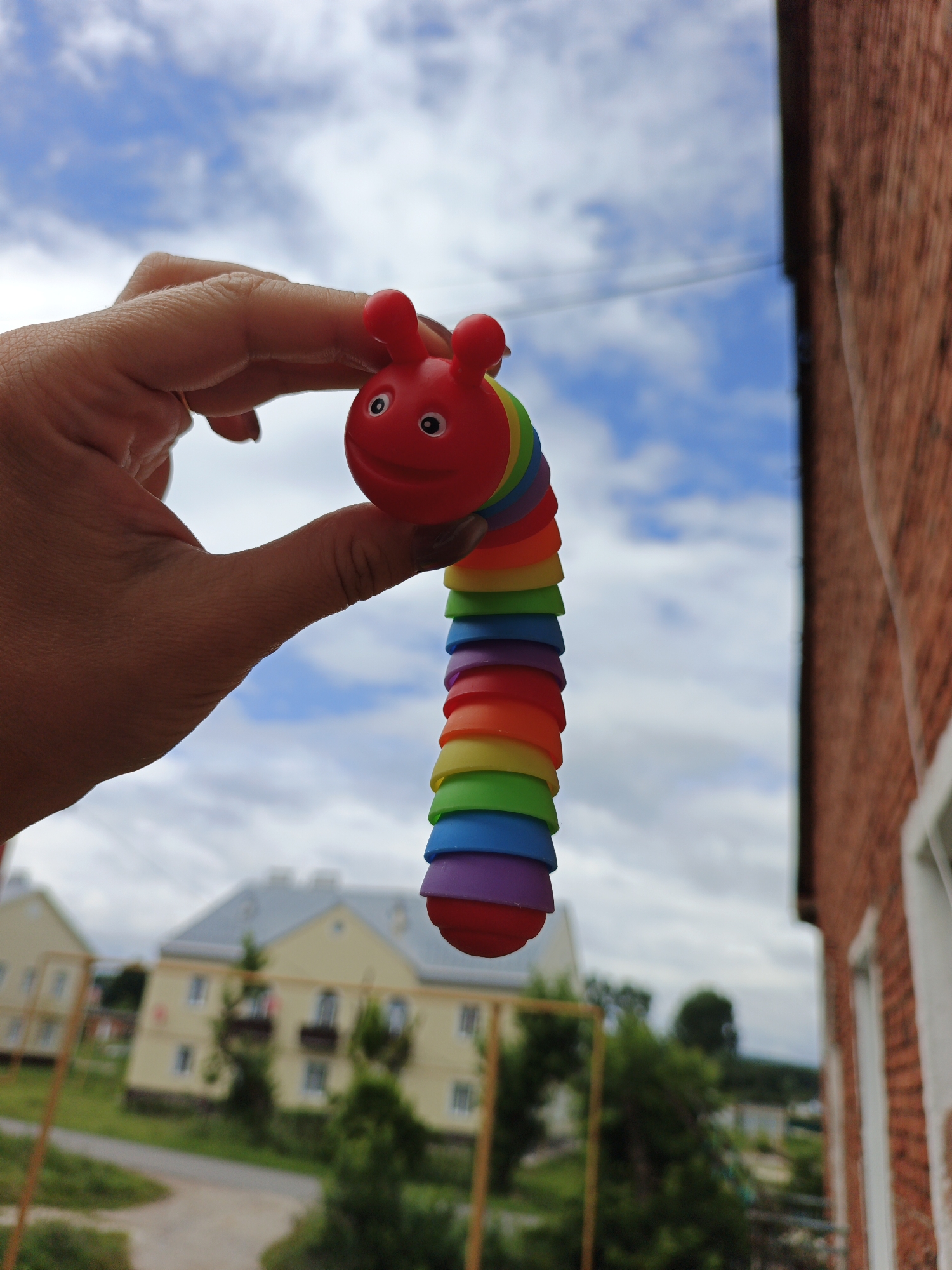 A person's hand is holding a rainbow-colored caterpillar toy with a red head and googly eyes against a cloudy blue sky. The toy is positioned in the foreground, with its body extending downwards in a segmented, curved fashion. In the background, out of focus, are yellow buildings with white trim and gray roofs, suggesting a residential area. A red brick building is visible on the right side of the image. The scene is set in the city of Medvedyovka, Russia.