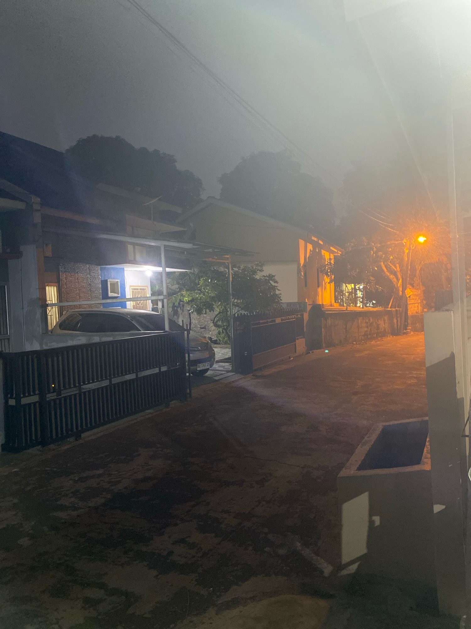 The image depicts a dark, hazy outdoor scene at night, likely in Palembang, Indonesia, as suggested by the license plate BG 2343 ZY which is characteristic of South Sumatra. A white car is parked in front of a house, partially obscured by a black fence. The house has a blue and white facade and is illuminated by artificial lights. To the right, a street lamp casts an orange glow on a paved area with a stone wall and vegetation. The overall atmosphere is subdued, with low visibility due to the haze or fog. The scene appears to be a residential area.