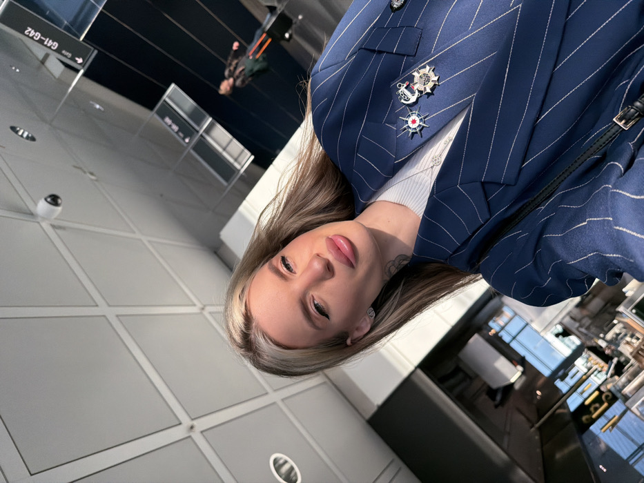 The image captures an upward-tilted selfie of a blonde woman located in a modern airport terminal. The woman, positioned in the foreground, has long blonde hair, prominent lips, and visible eyeliner, with a small tattoo present on her left neck. She wears a navy blue pinstriped blazer over a white ribbed top and a silver necklace. The blazer is embellished with three distinct pins: a red and white star-like emblem, a Russian naval flag-like emblem, and a multi-pointed star-shaped medal. A white dome security camera is mounted on the paneled ceiling overhead. In the mid-ground, a black sign with white lettering explicitly displays "Gate G41-G42." A blurry figure of another individual pulling luggage is visible in the distant background. The location is identified as Munich Airport (MUC), which is situated in Freising, Germany.