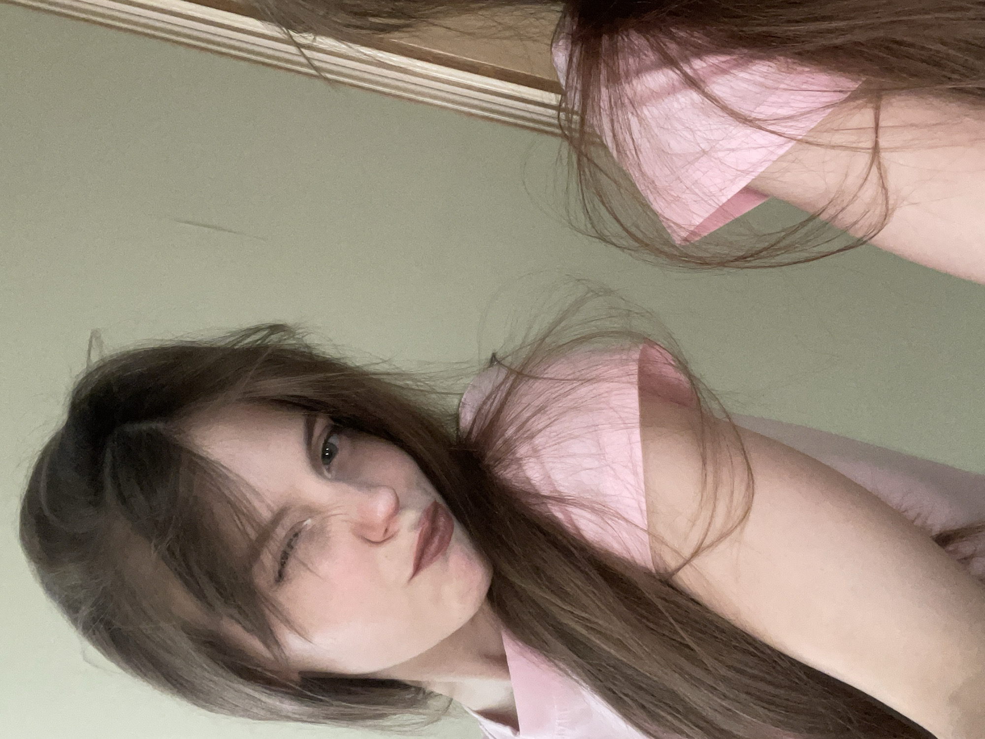 A selfie of a young woman with light skin, brown hair, and light green eyes is taken indoors. She is winking her left eye and has a slight frown. She is wearing a light pink collared shirt. Part of her reflection is visible in what appears to be a mirror above her. The wall behind her is a pale green color with a light wood trim along the top. The location cannot be determined.