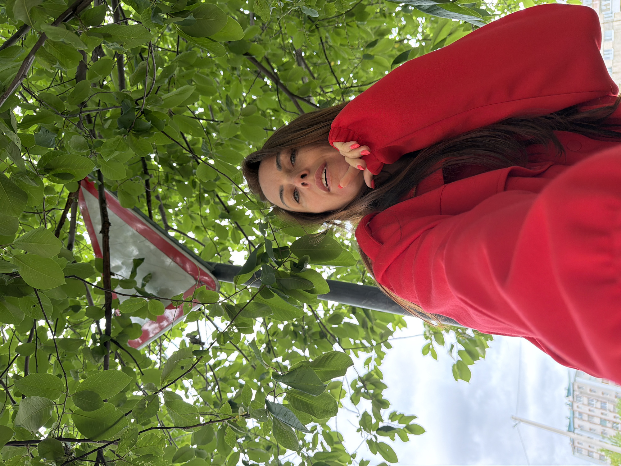 A low-angle selfie captures a woman outdoors in Birobidzhan, Russia, under an overhead canopy of dense green leaves and branches. The woman, with long brown hair, wears a bright red long-sleeved top or jacket. Her right hand, adorned with bright pink painted nails, is positioned near her chin, with fingers slightly curled. Partially visible through the foliage, above and to the right of the woman, is an inverted white triangular road sign with a prominent red border, consistent with a "Give Way" or "Yield" sign, affixed to a dark pole. The sky in the background is light and overcast. In the distant lower-left corner, faint outlines of light-colored multi-story buildings and utility poles are discernible.