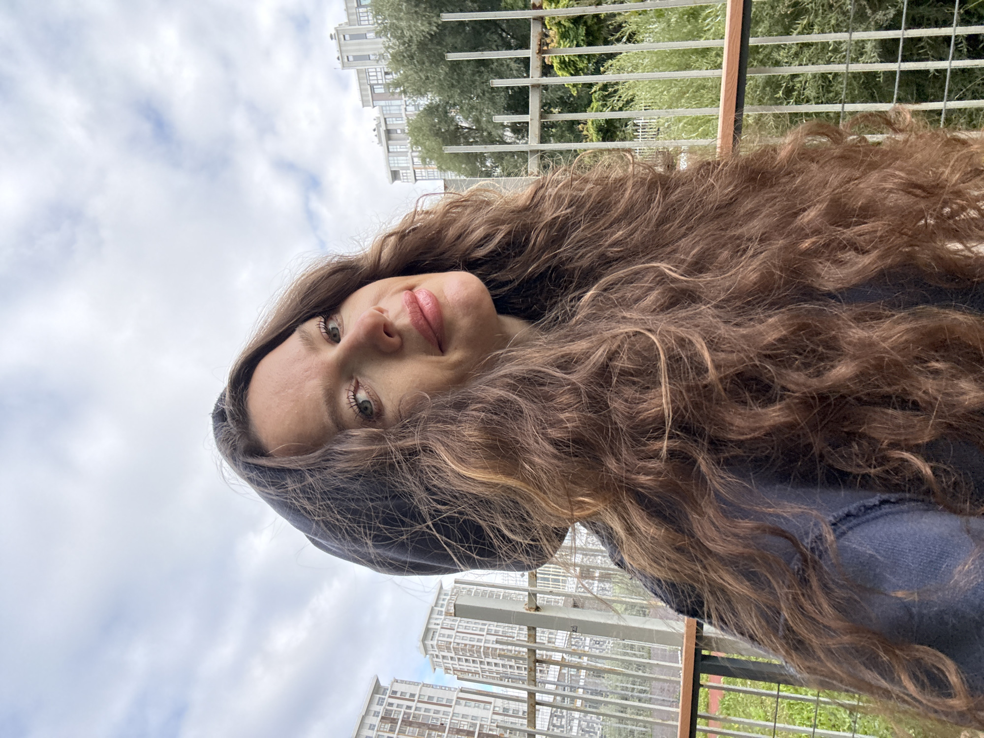 A medium shot from a low angle shows a woman with long, curly brown hair, wearing a dark hooded sweatshirt. She is looking slightly off-camera to her right with a subtle smile. In the background, there are several tall, modern apartment buildings, a metal fence, and lush green trees. The sky is partly cloudy.