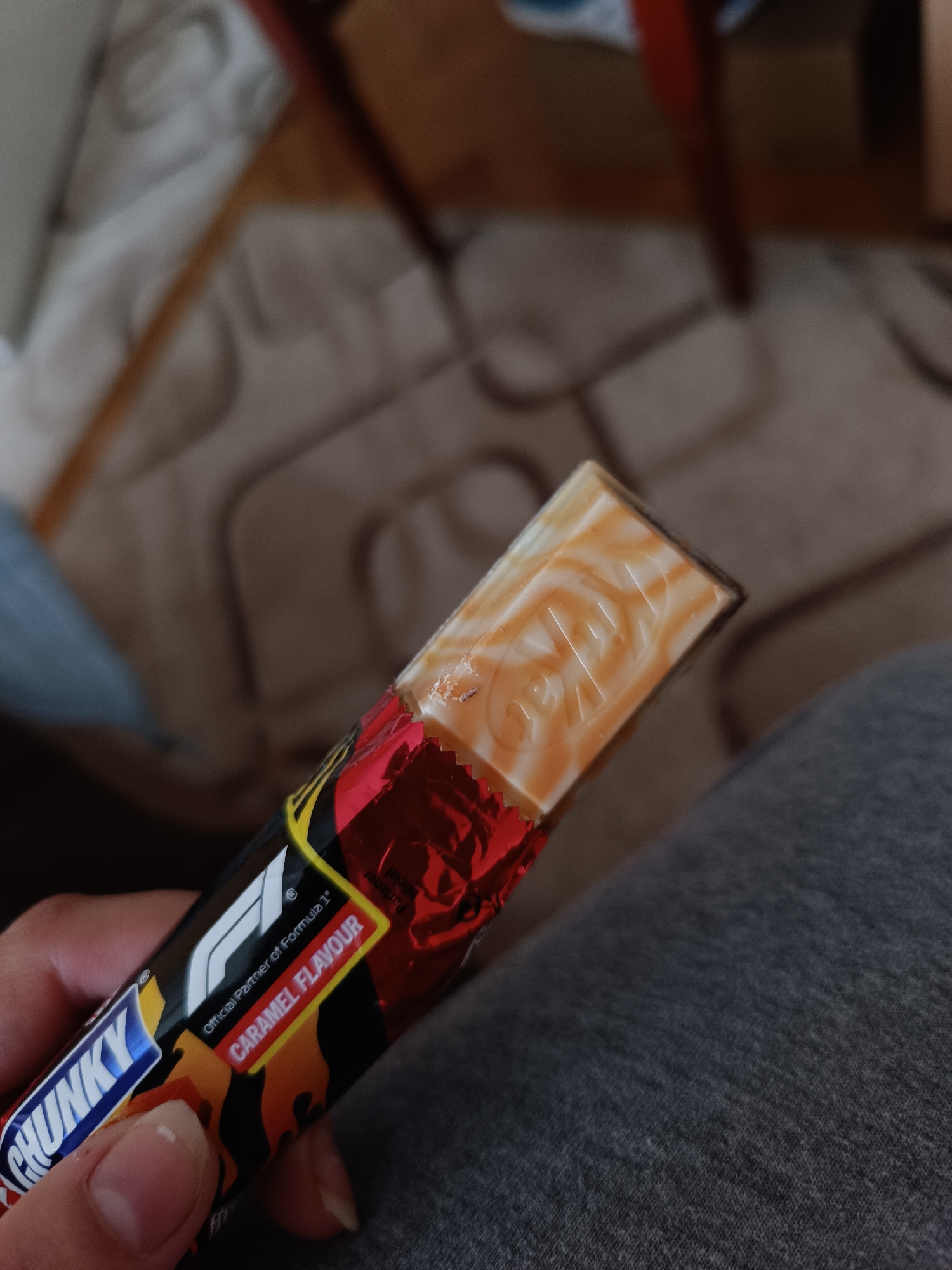 A close-up shot captures a person's hand holding a partially opened KitKat Chunky chocolate bar in an indoor setting in Pleven, Bulgaria. The hand, with visible fingers and neatly kept nails, grasps the red and black wrapper of the confection.
The chocolate bar is clearly a "KitKat CHUNKY" with a "CARAMEL FLAVOUR," as indicated by the text on its packaging. It proudly displays the "F1" logo and states "Official Partner of Formula 1®," suggesting a special edition product. The top portion of the chocolate bar is exposed, revealing a surface with intricate swirls of white and caramel-colored coating, typical of a caramel-flavored chocolate. The brand name "KitKat" is subtly embossed into this caramel layer.
The background is softly blurred, indicating a shallow depth of field, and shows elements of a domestic environment. A light-colored patterned rug or carpet is visible, along with the dark, possibly wooden, legs of furniture like a chair or table. To the right, a segment of grey fabric, likely clothing such as a pant leg or a blanket, is present. The lighting is consistent with an indoor setting, but the specific time of day is not discernible. No particular activity beyond holding the chocolate is depicted, and no specific emotions are conveyed.