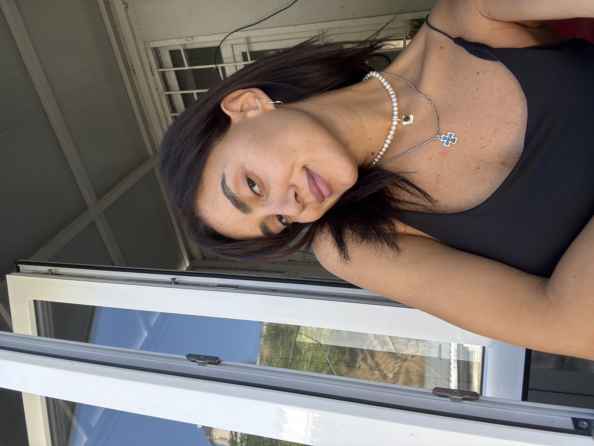 A woman with dark hair and medium skin tone, wearing a black tank top and two necklaces, one of pearls and one with a cross pendant, is positioned in front of an open window. The window frames a view of a hilly landscape with buildings and greenery. The woman is looking towards the camera and smiling slightly. The lighting suggests it is daytime, possibly late afternoon due to the warm tones. The overall setting appears to be a residential area, perhaps in a city given the presence of buildings in the background. The style of the window and the architectural details of the background building suggest a European location, possibly Eastern Europe or the Caucasus region.