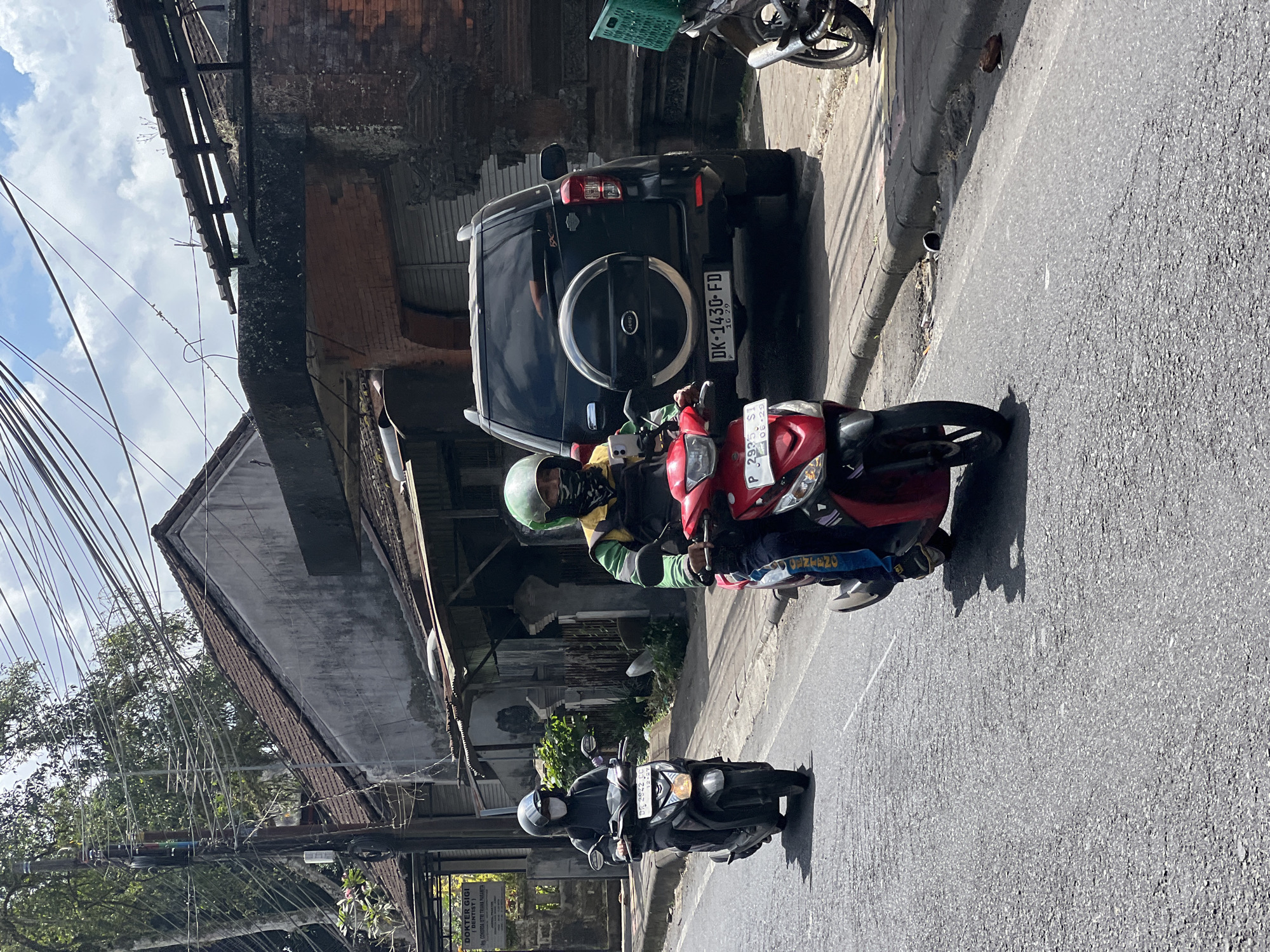 In Payangan, Indonesia, two individuals are seen riding motorcycles on a paved road on a sunny day. The person in the foreground, on a red scooter, is wearing a green helmet, a yellow and green jacket, and a patterned bandana covering their face. They are looking at their phone, which is mounted on the handlebars. Behind them, on a dark grey scooter, another person is wearing a light grey helmet, a dark grey hoodie, and a white mask. They are also wearing a backpack.
The road is lined with buildings and vegetation. To the right, a black SUV with a spare tire mounted on the back is parked. The license plate reads "DK-1430-FD". The buildings appear to be a mix of traditional and modern architecture, with tiled roofs and brick walls. Overhead, a dense network of electrical wires stretches across the sky. In the background, lush green trees suggest the presence of a tropical environment. The overall scene depicts a typical street view in a bustling Indonesian town.