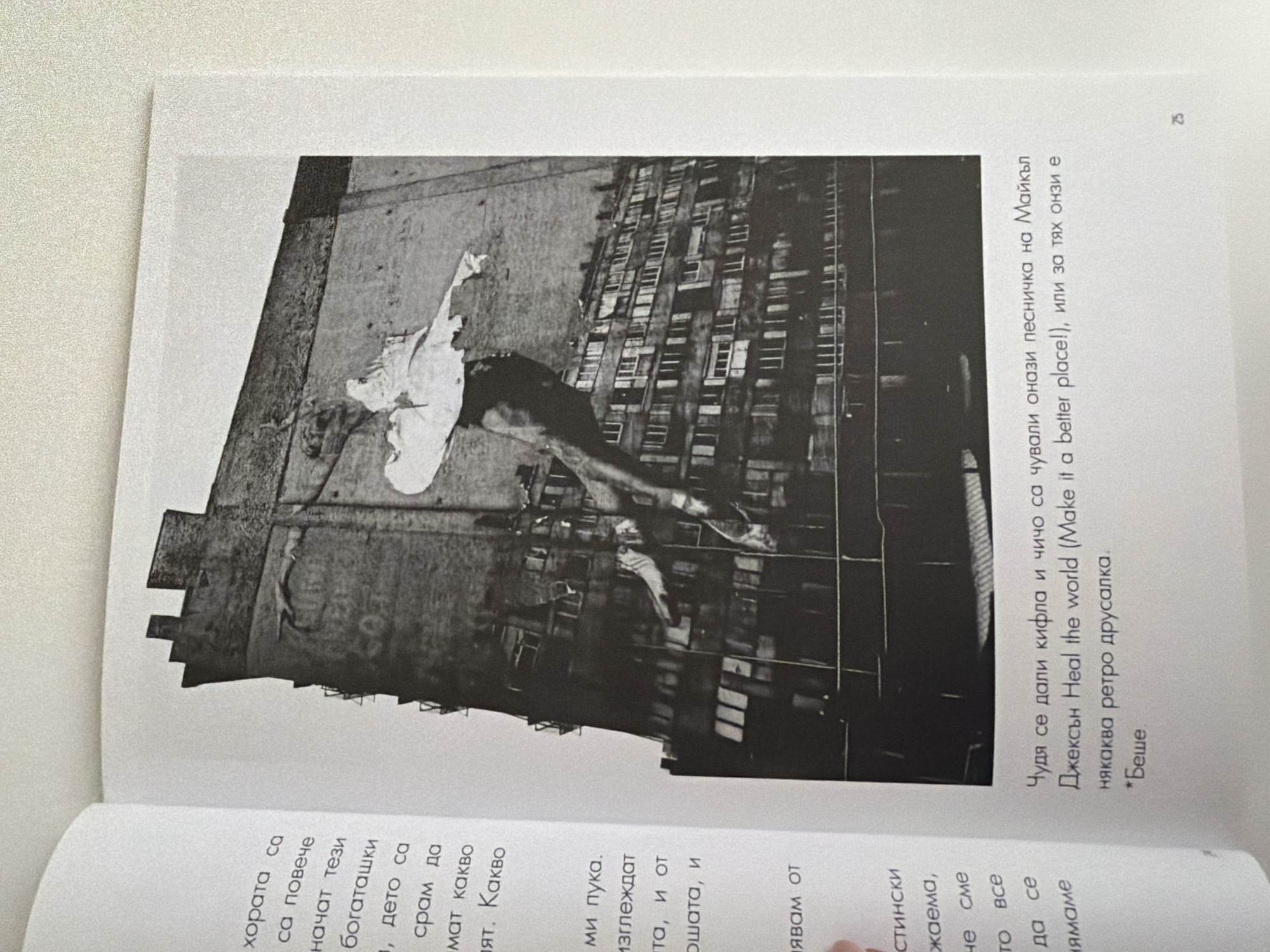 The image displays an open book with text on the left page and a black and white photograph on the right. The photograph shows a large, somewhat dilapidated building with many windows, resembling an apartment complex, and a dark, textured facade. Superimposed on the building is an image of a figure in a ballet pose, appearing to be a dancer, leaping or floating against the building's surface. The figure seems to be wearing a light-colored outfit. There is text printed below the photograph, which appears to be in Bulgarian. On the far right edge of the page, a small number "25" is visible, likely indicating a page number. The text on the left page is also in Bulgarian.