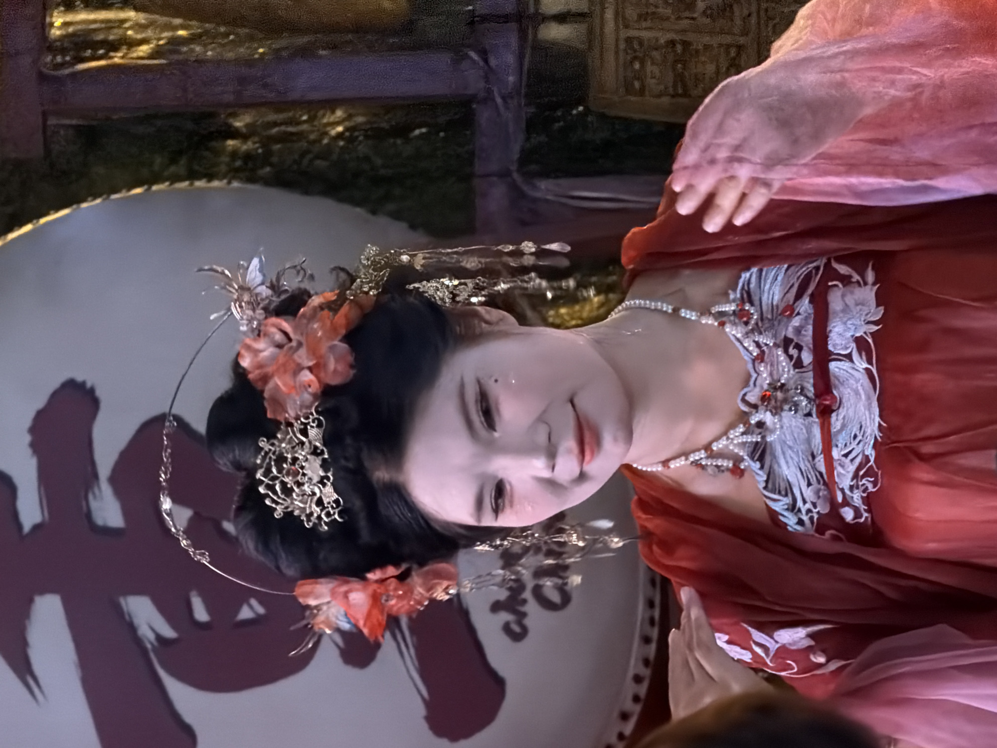 The image shows a close-up of a woman in traditional Chinese attire. She has black hair styled in an updo, adorned with ornate golden hairpins and red flowers. Her face is painted with white foundation, and she has subtle eye makeup and lipstick. She is wearing a red garment with white embroidery and a pearl necklace with red accents. Her left shoulder is touched by another person's hand, whose arm is visible in the lower left corner. Behind her, a large, dark red Chinese character is painted on a light-colored surface. The background to the right appears to be a dark, textured wall with some golden elements. The overall scene suggests a theatrical performance or a cultural event.