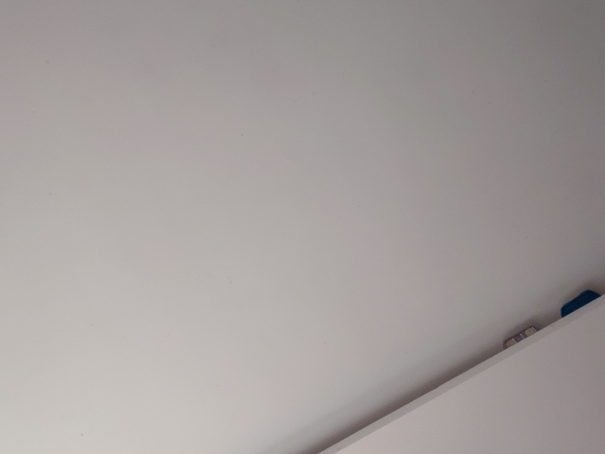 The image presents a close-up, static view of an indoor environment, dominated by a plain, light gray or off-white surface, likely a wall or ceiling. This surface fills most of the frame and appears subtly textured with minor specks.
In the bottom right corner, a distinct, straight white line cuts diagonally across, marking the edge of another architectural feature, such as a shelf, partition, or a cabinet. Behind this edge, several small objects are partially visible. There are two or three small, metallic, rectangular items, possibly screws, electrical connectors, or decorative elements, reflecting a faint gleam. To the immediate right of these metallic objects, a small, dark blue, block-like item, possibly made of plastic or fabric, is tucked into the narrow space, partially hidden by the white edge.
The lighting is soft and even, suggesting either diffused natural light from an unseen window or ambient artificial illumination. There are no harsh shadows. No people or specific activities are depicted. The scene is quiet and minimalist, focusing on structural details and small, overlooked objects within an interior space. There is no visible text. The image does not contain any specific visual cues to indicate its location in Santa Ana, Venezuela.