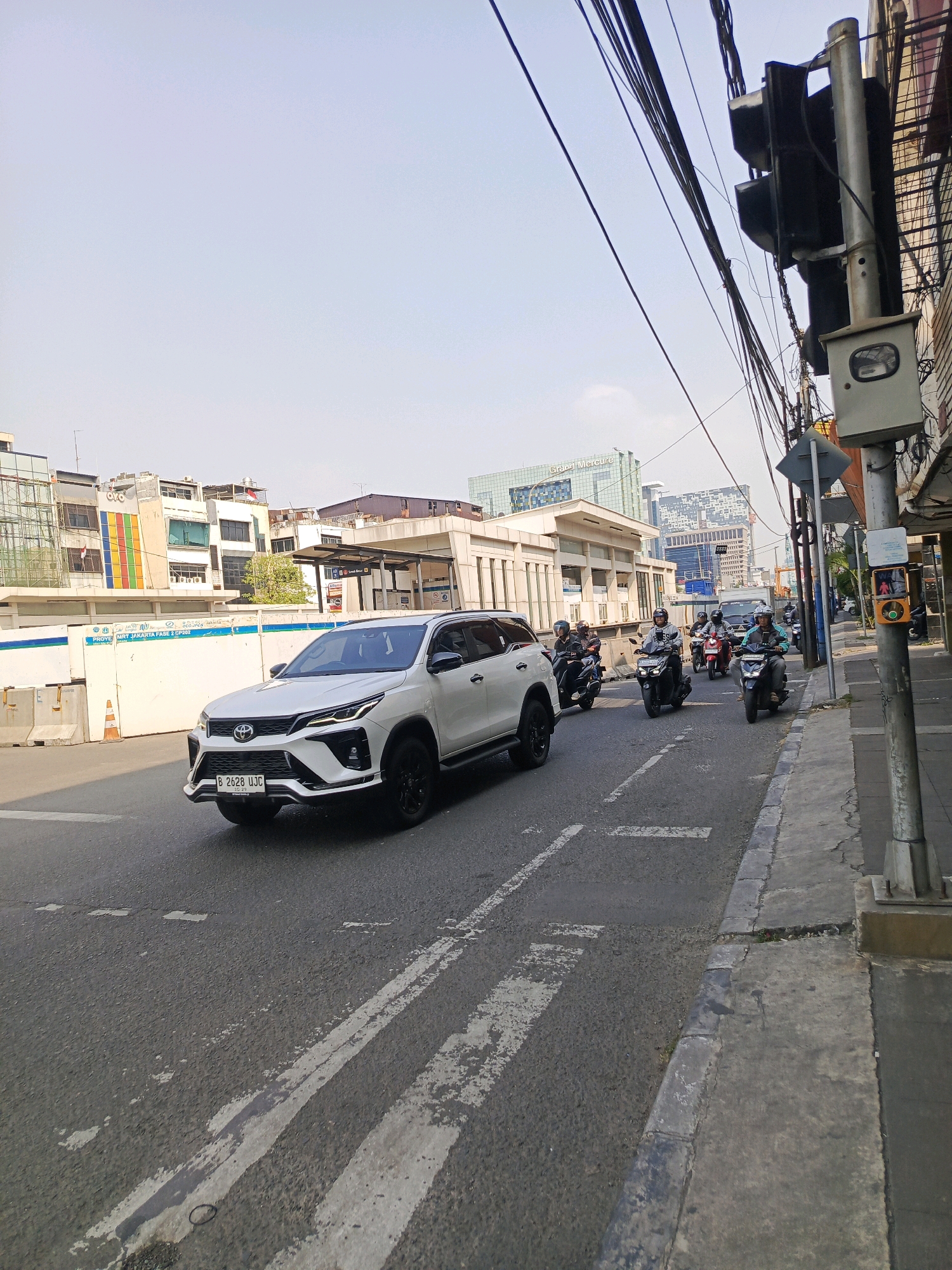 In West Jakarta, Indonesia, a white Toyota Fortuner SUV, license plate B 2628 UJC, is captured in motion on a city street. The vehicle is in the left lane of a two-lane road, heading away from the camera. Several motorcyclists are also on the road, some in front of and beside the SUV, all appearing to be moving in the same direction. The road is marked with white dashed lines, indicating lanes.
On the right side of the street, a sidewalk is visible, bordered by a curb. A utility pole with traffic signals and electrical wires is prominently positioned on the sidewalk. A pedestrian crossing signal, showing a red standing figure, is attached to the pole. Adjacent to the pole is a grey traffic camera box.
In the background, buildings of various sizes and architectural styles line the street, suggesting an urban environment. Construction fencing is visible on the left side, with concrete barriers and an orange traffic cone. Power lines crisscross the sky, a common sight in dense urban areas. The sky is clear and appears to be a bright, hazy blue, indicating daytime. The overall scene depicts typical street traffic and urban infrastructure in a city setting.