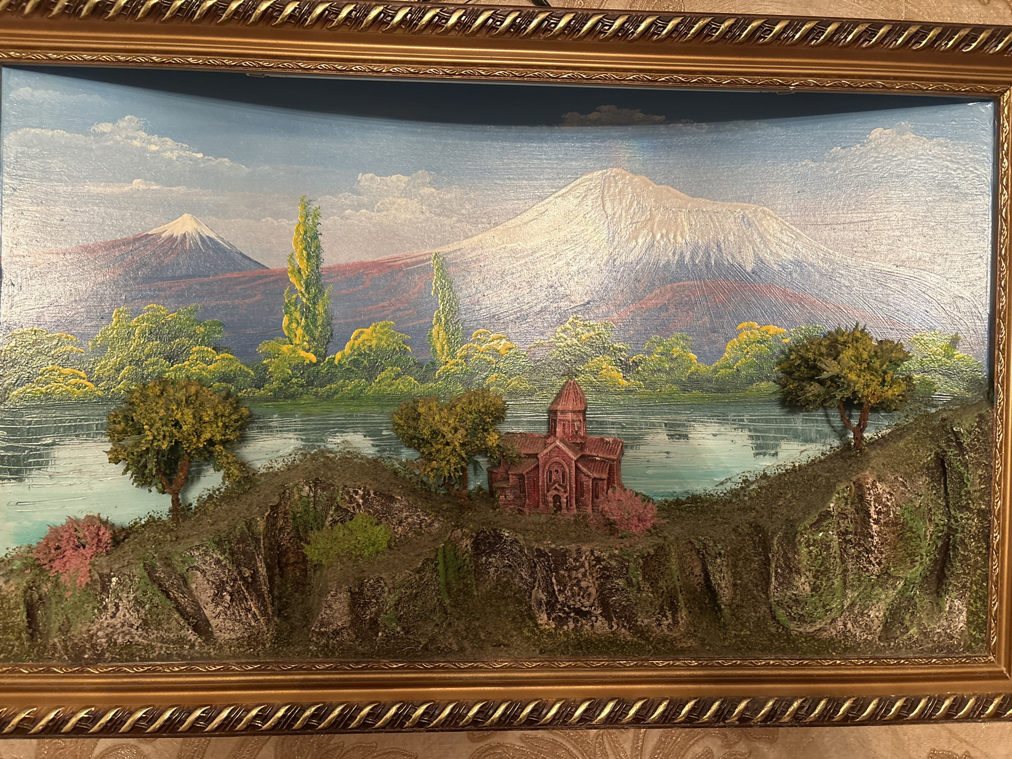 The image depicts a framed painting of a landscape featuring mountains, a lake, trees, and a church-like structure. Two prominent mountains are visible in the background, with one appearing to be snow-capped. The landscape includes various shades of green vegetation and what looks to be a body of water. A red, church-like building sits amongst the trees and terrain. The painting is encased in an ornate gold-colored frame. There is no indication of the painting's location in Krasnoyarsk, Russia, or any other city. No people or actions are depicted.