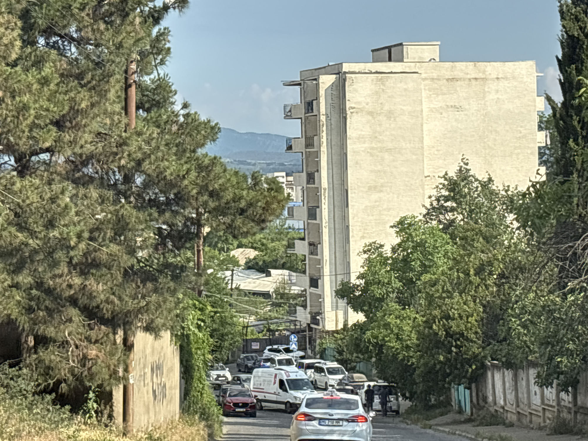 The image depicts a street scene in Tbilisi, Georgia. A tall, light-colored apartment building with multiple balconies dominates the background on the right side of the frame. The building's facade appears somewhat weathered. In the foreground, a road slopes downwards and is lined with trees and vegetation on both sides. There is a significant amount of traffic on the road, consisting of various cars, vans, and what appears to be a taxi. Several people are visible, some standing near vehicles and others potentially driving. The overall environment suggests a residential area within the city, with a mix of urban infrastructure and natural elements. The sky is clear and blue.