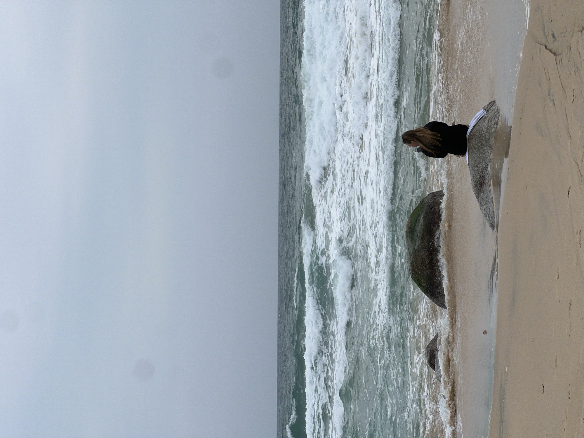 A lone woman with long blonde hair is seated on a large, flat rock on the sandy shore of a beach. She is wearing a dark-colored hooded jacket and appears to be holding a small object, possibly a phone or camera, to her face. The rock she is sitting on is partially submerged in shallow water where waves are breaking. To her left, two smaller rocks are visible in the surf, and a larger, moss-covered rock is situated further to the left, closer to the water's edge. The ocean exhibits moderate waves with whitecaps, and the sky is overcast with a grayish hue. The sand is light brown and shows subtle textures and water lines. The scene suggests a peaceful yet somewhat melancholic atmosphere, potentially during an off-season period given the overcast conditions and absence of other beachgoers. The location is the beach in Choeng Thale, Thailand.