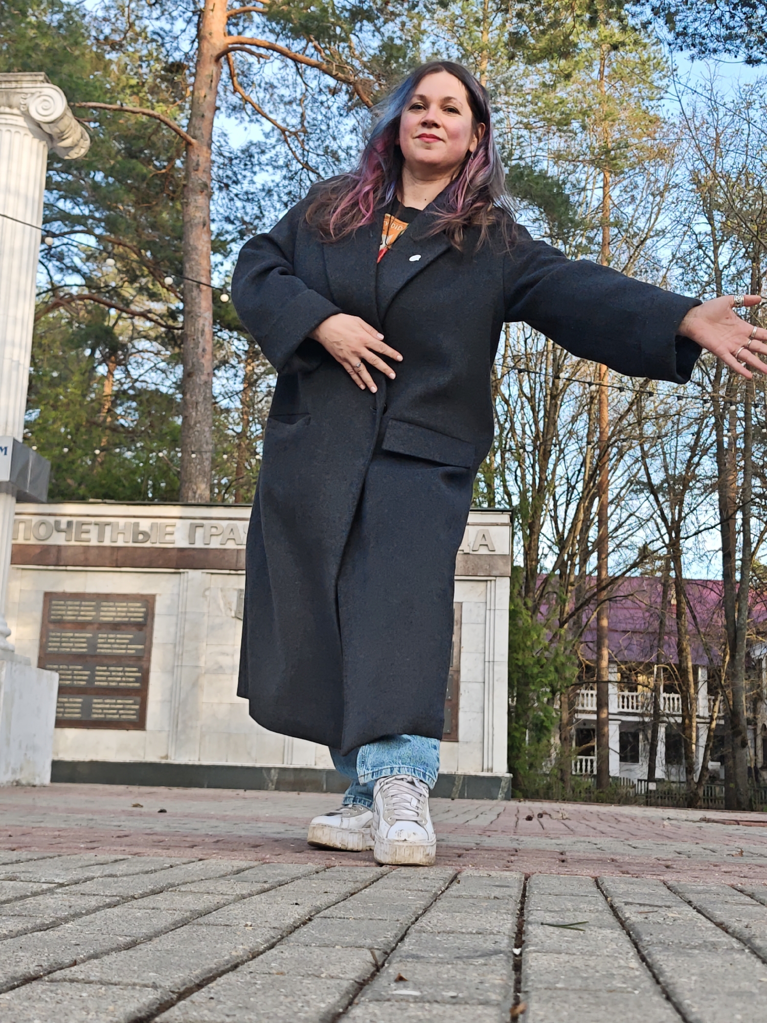 A woman with pink-streaked hair, wearing a long grey coat and jeans, stands on a brick-paved area. She has one hand on her chest and the other extended outward. She is wearing white sneakers. Behind her is a monument with the inscription "ПОЧЕТНЫЕ ГРА" written in Cyrillic. Trees are visible in the background, including a large pine tree, and a white building with a purple roof is partially visible. The location is consistent with a public space, possibly a park or plaza, in Krasnaya Gorka, Russia.
