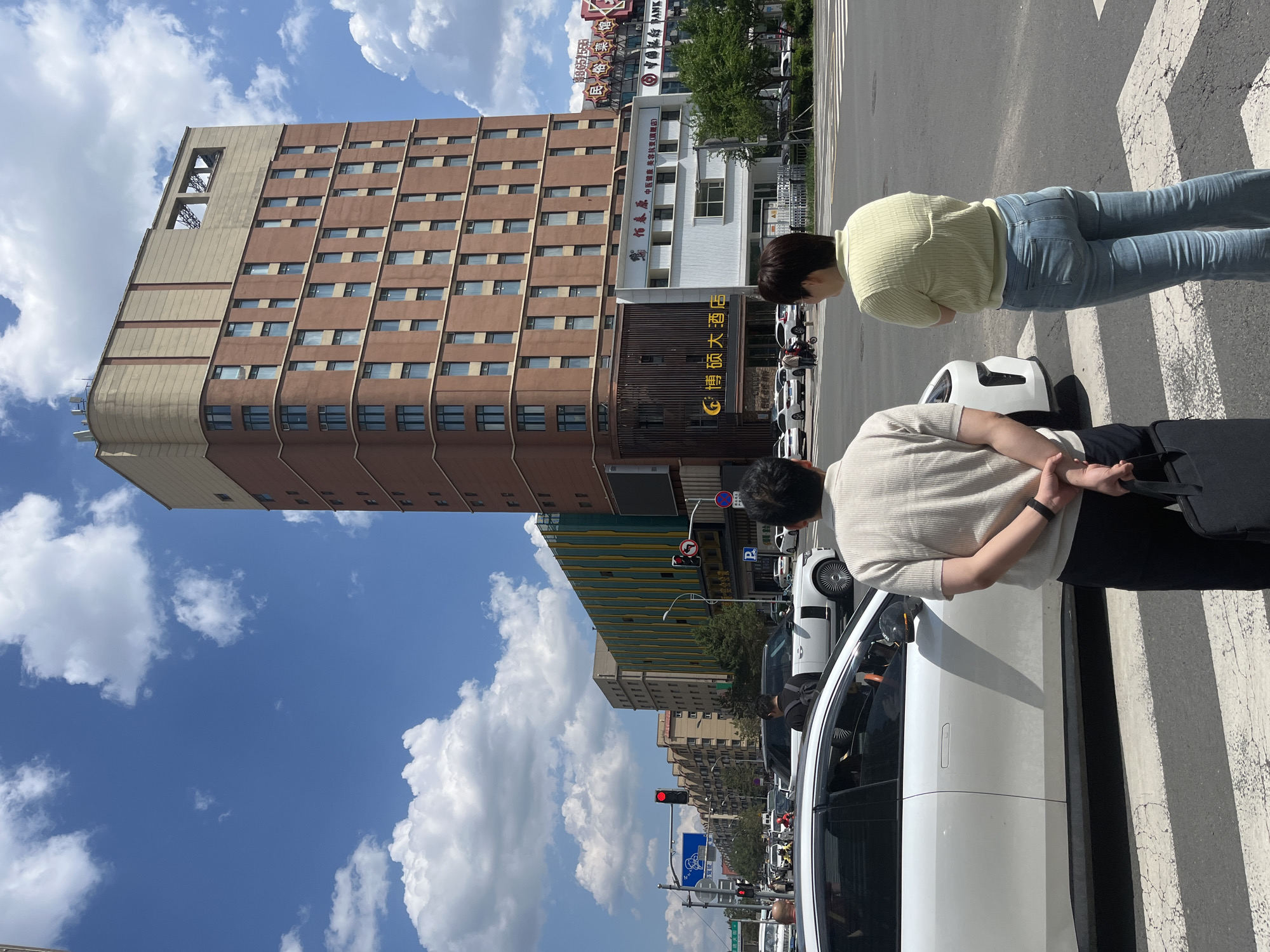 The image shows a street scene in Changchun City, China. Two people stand on a crosswalk next to a white car. A tall, brown building stands in the background, along with other buildings and street signs. The sky is blue with white clouds. There are many vehicles on the street.