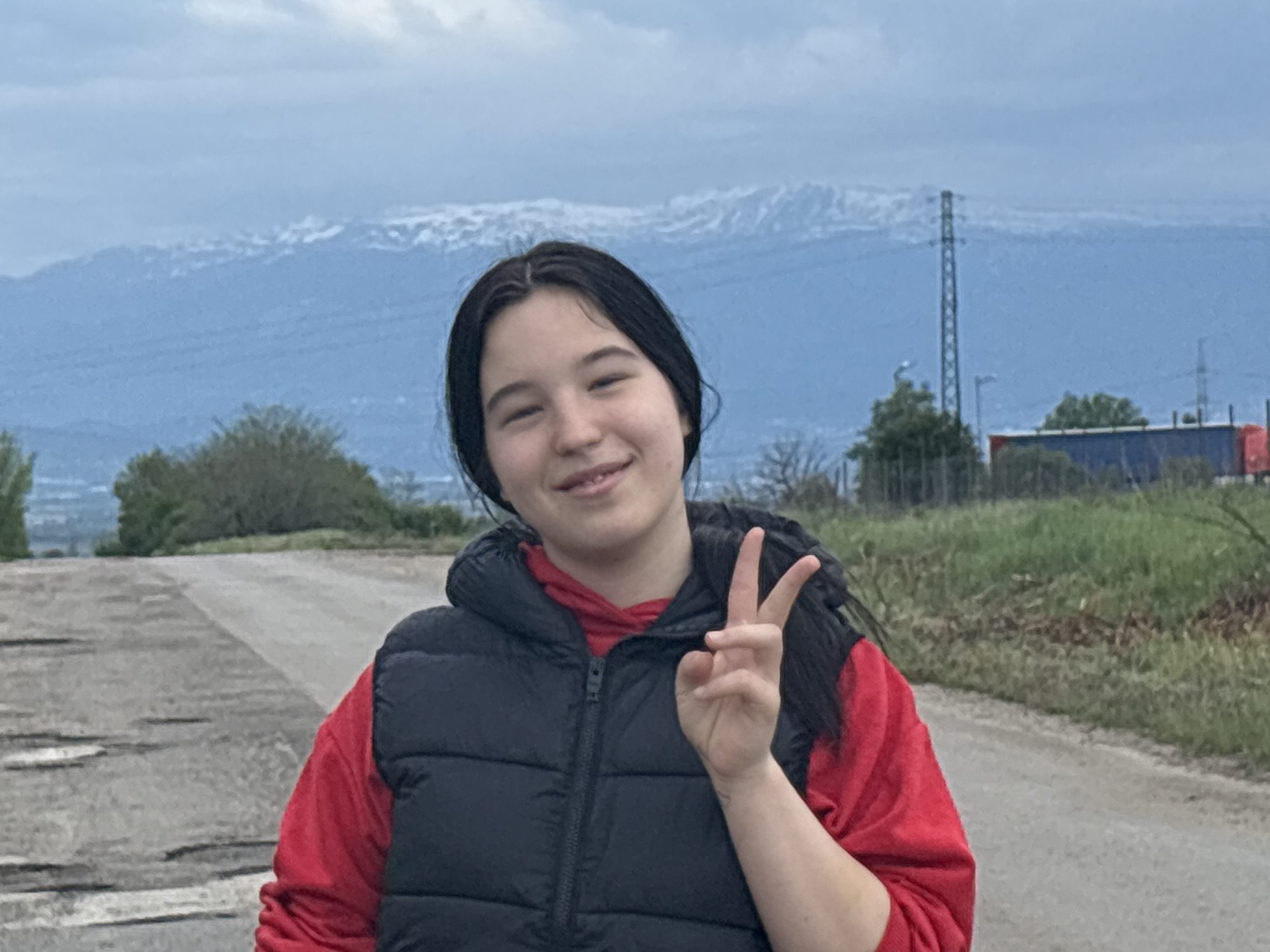 A young woman stands on a paved road in Eleshnitsa, Bulgaria, making a peace sign with her right hand. She wears a red hooded sweatshirt under a black puffer vest. The road is cracked and worn. Behind her, a landscape of sparse vegetation transitions into a backdrop of distant mountains capped with snow. A utility pole and a glimpse of a blue and red trailer are visible on the right side of the frame. The sky is overcast.