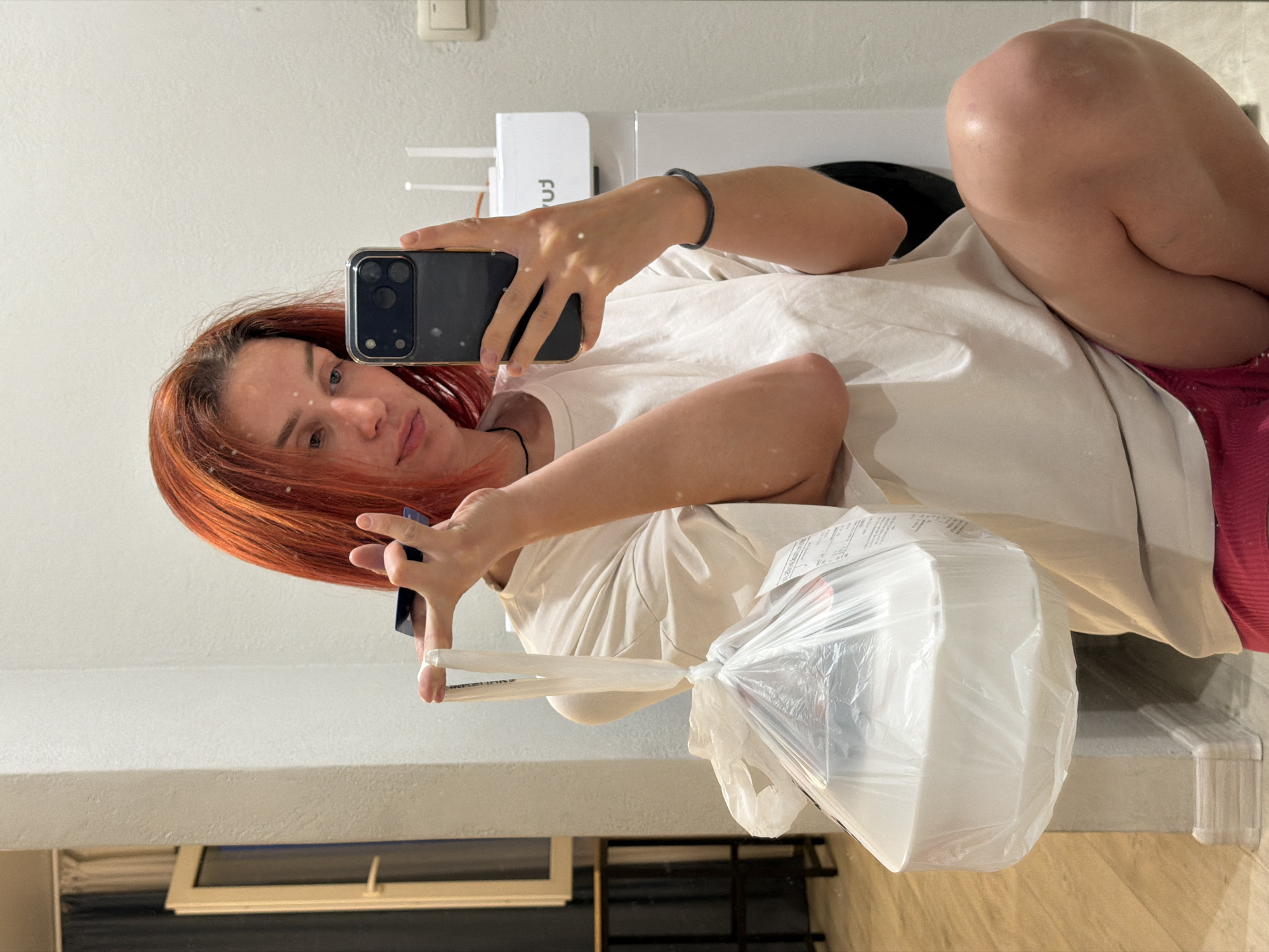 A woman with red hair is taking a selfie in what appears to be a bathroom. She's holding a smartphone in her left hand and a plastic bag filled with white containers in her right. She's wearing a light-colored t-shirt and pink shorts. The background includes a toilet, a white wall, and a window. The location is Naberezhnye Chelny, Russia, inferred from details in the background which is not visible in crops.