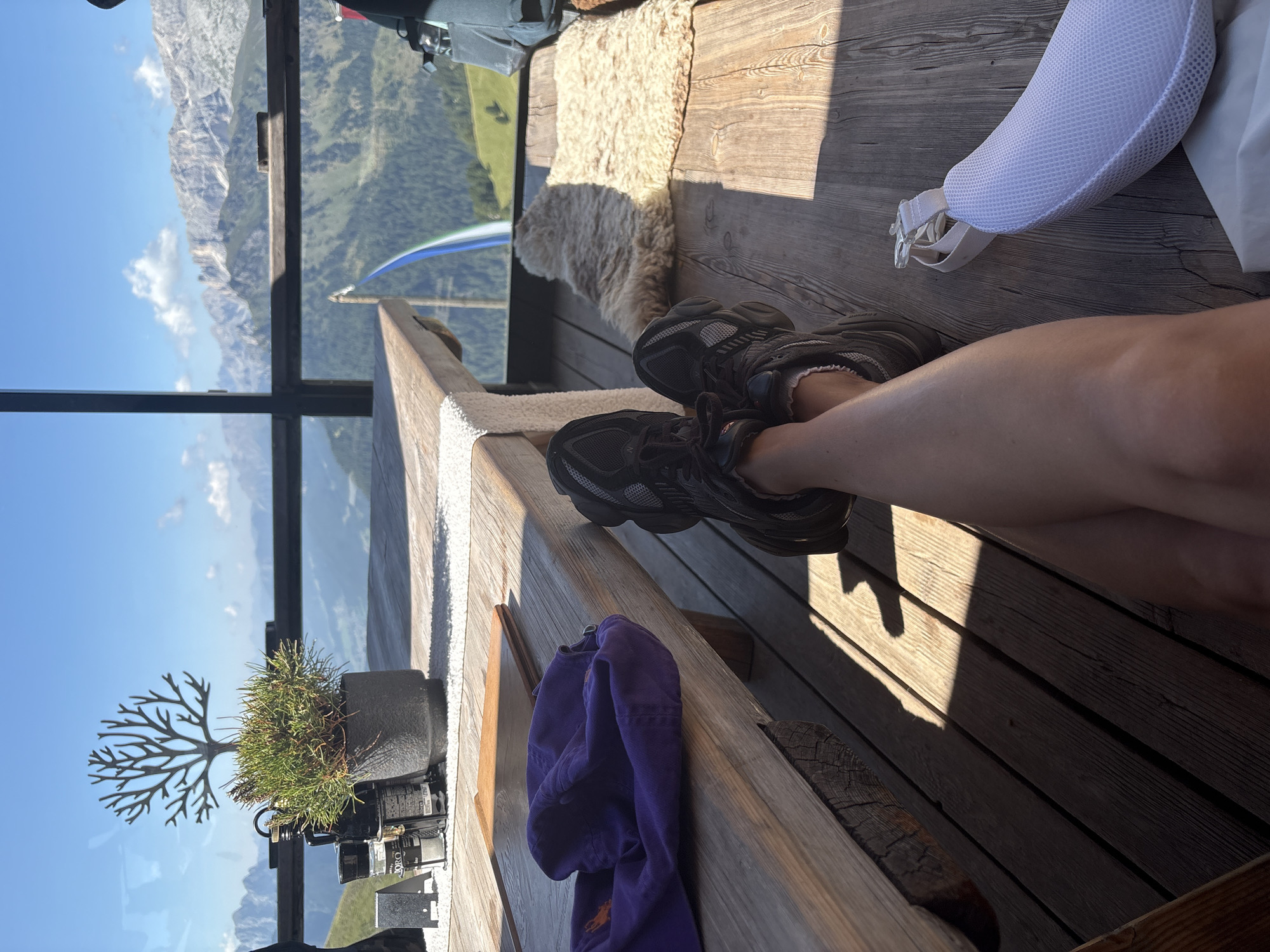 A first-person perspective shot shows a person's legs crossed and resting on a wooden deck. They are wearing dark athletic shoes and white ankle socks. In the foreground, a purple baseball cap lies on a wooden table. To the right of the person's legs, a light-colored, furry rug is placed on the deck, and a white mesh cushion is visible. Beyond the deck, a panoramic view of majestic mountains under a clear blue sky with scattered clouds is visible, suggesting a location in Campitello di Fassa, Italy, likely a restaurant or viewing platform offering a scenic vista. The environment is sunny with strong shadows cast by the table and railing.