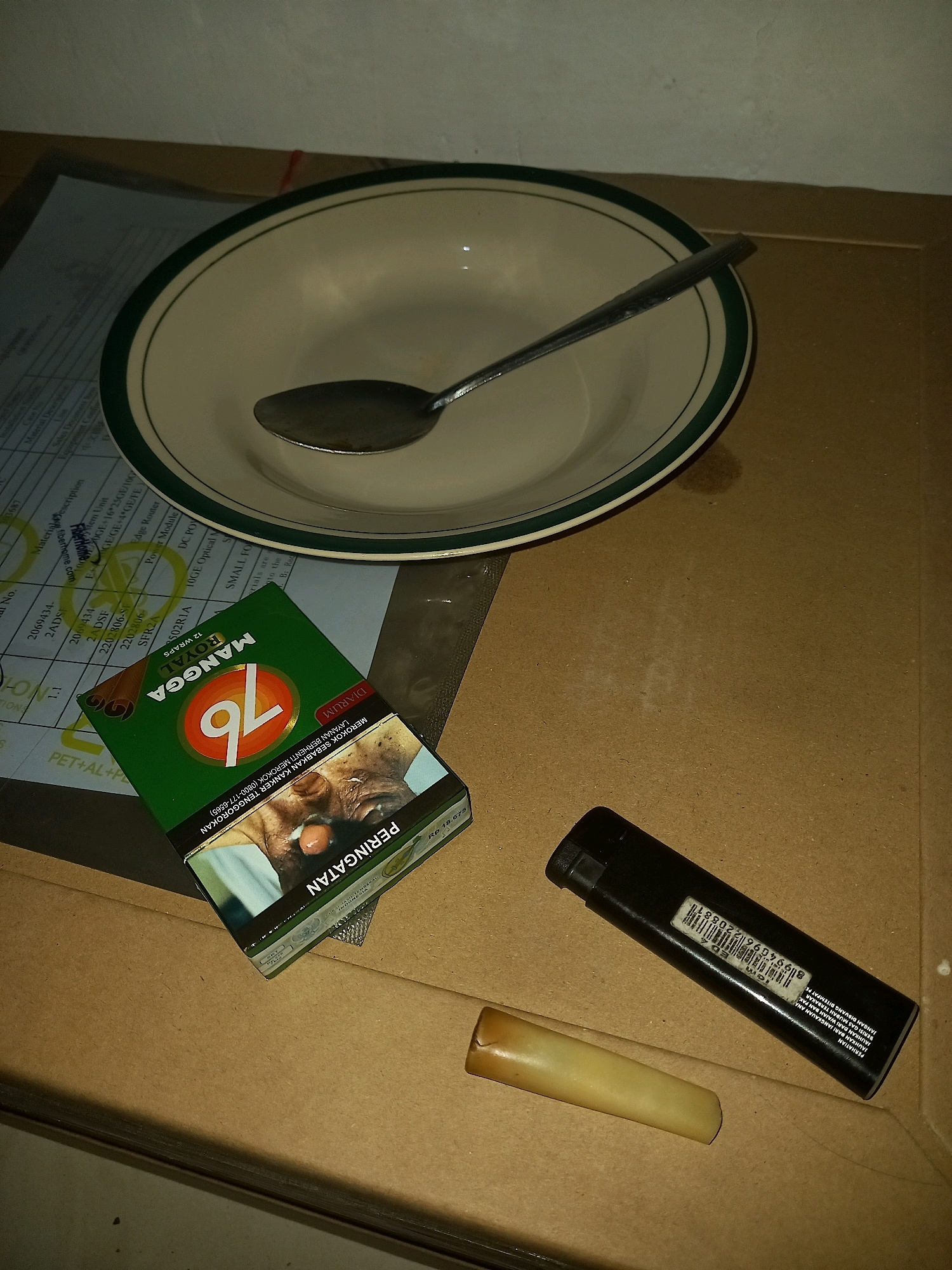A close-up, overhead shot shows a collection of objects on a brown, cardboard surface. In the upper left quadrant is a white ceramic bowl with a dark green rim, containing a silver spoon. To the left of the bowl is a document with text and a grid. In the lower left quadrant is a green cigarette pack labeled "MANGO ROYAL 76" with a warning label featuring an image of a person's face. To the right of the cigarette pack are two objects: a black lighter and a light brown, cylindrical object with a tapered end. The scene appears to be indoors, possibly in Mejasem Timur, Indonesia, based on the text on the cigarette pack, which includes Indonesian language warnings. The lighting is dim, casting shadows and creating a slightly muted color palette. The overall impression is one of everyday objects arranged casually.