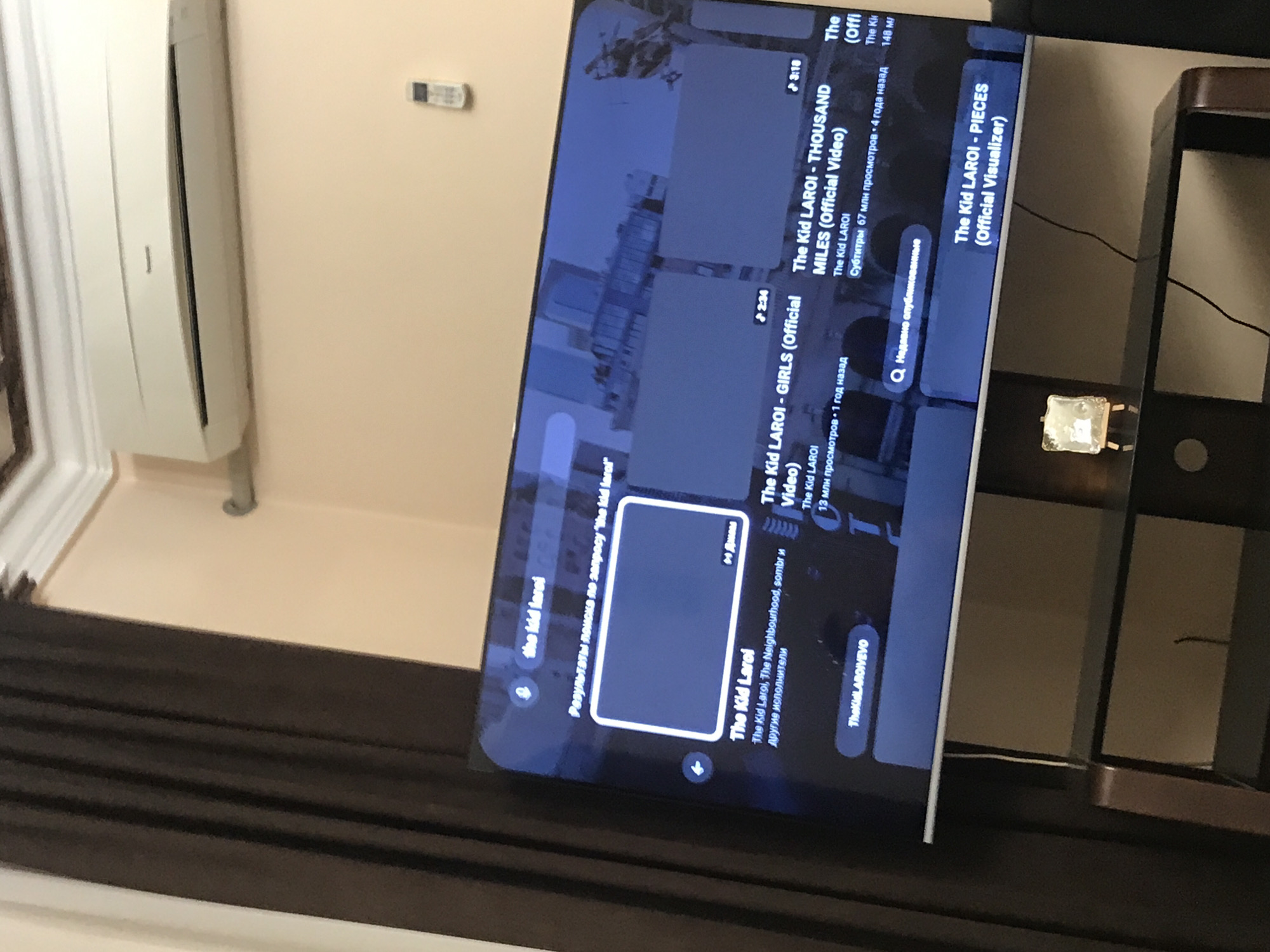 The image shows a television screen displaying a YouTube search result for "the kid laroi." The results include songs by The Kid LAROI such as "GIRLS (official video)," "THOUSAND MILES (official video)," and "PIECES (official Visualizer)." The television is placed on a glass and dark-colored wooden stand. In the background, an air conditioner is mounted on a beige wall. A dark curtain is partially visible in the foreground. The location in the city of Yerevan, Armenia, is not discernible from this image.