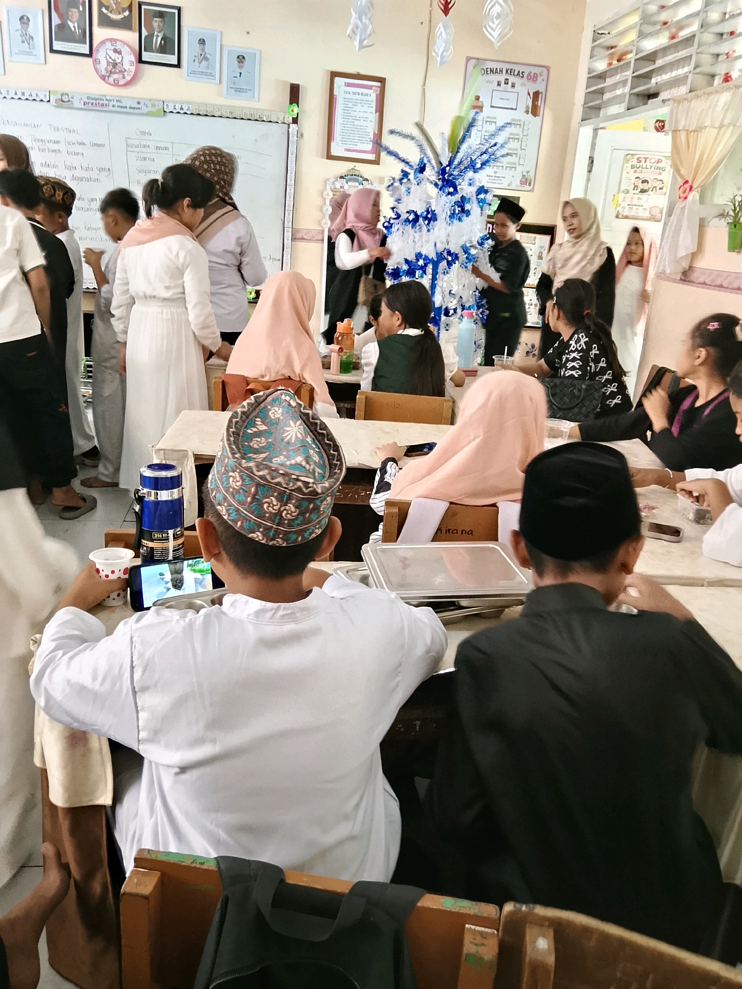The image depicts a classroom setting, likely in Besusu Timur, Indonesia, filled with students and adults. The room appears to be decorated for an event, with a blue and white tiered structure resembling a Christmas tree in the center, adorned with paper cutouts. Students, many wearing traditional Indonesian attire like peci (caps) and hijabs, are seated at tables. One student in the foreground is looking at a mobile phone, while others are engaged in activities or conversations. The walls are adorned with framed pictures, educational charts, and decorations. A whiteboard with Indonesian writing is visible on the back wall. The environment suggests a lively, possibly festive, school event.