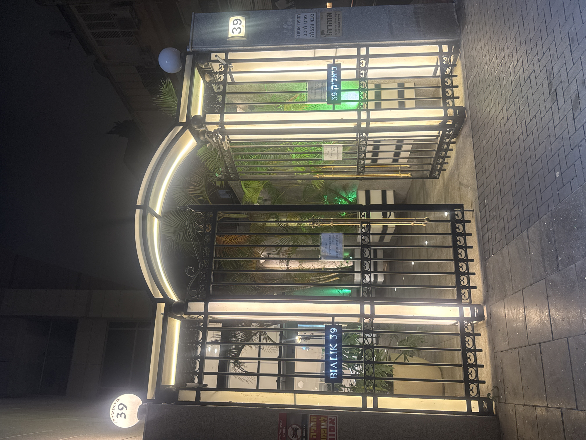 The image shows the entrance to 39 Bialik Street in Ramat Gan, Israel. The entrance has an arched gate with decorative ironwork, illuminated by lights. Signs indicate "Bialik 39" in Hebrew and English, as well as parking restrictions and a warning about closed-circuit television surveillance. Plants are visible behind the gate. The street appears to be paved with stone blocks. There are no people visible.