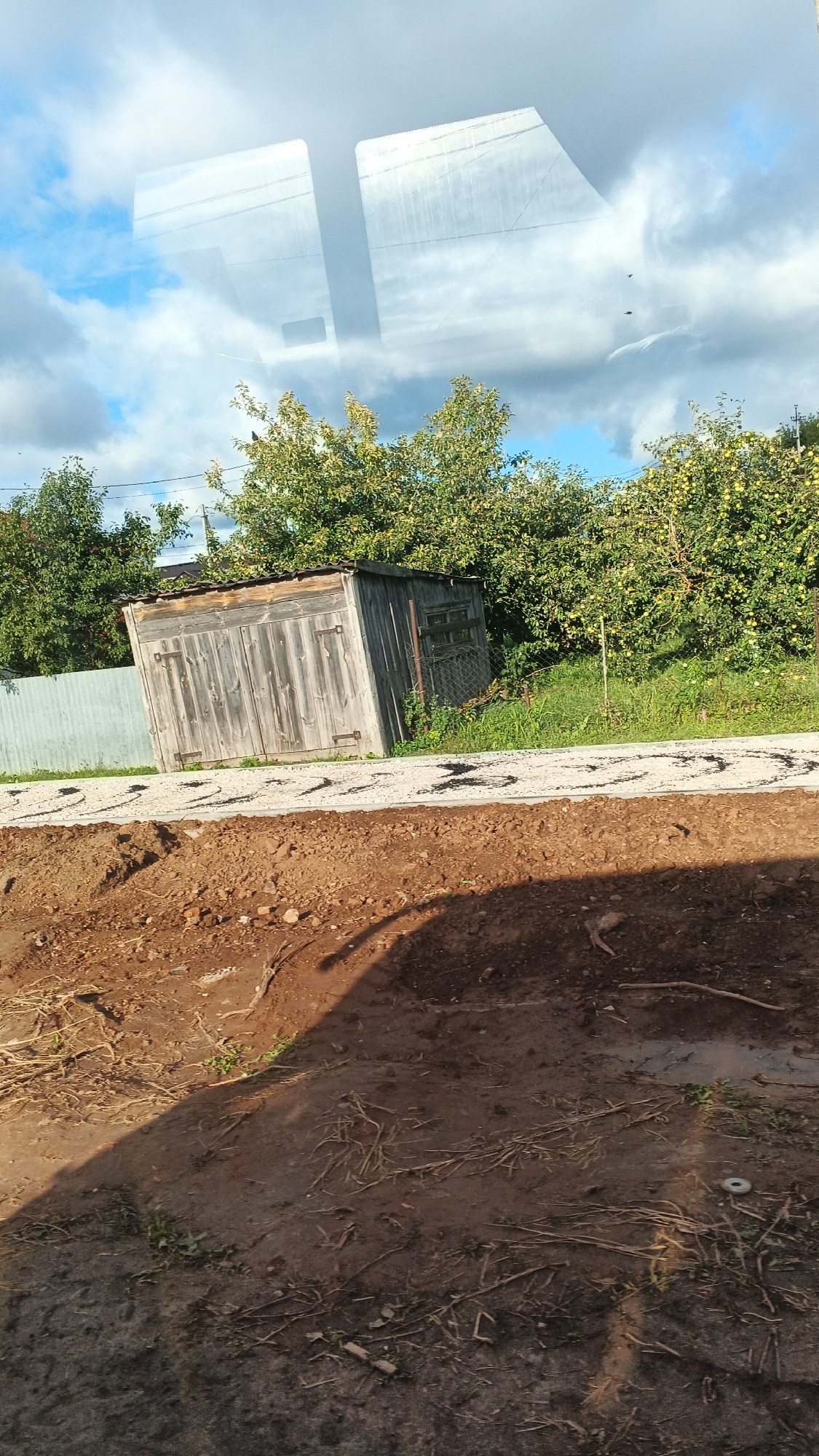 The image depicts an outdoor scene in Киясово, Russia. In the foreground, there is disturbed brown soil with scattered twigs and some small green plants. A dark shadow stretches across this area from the left. Behind the soil, a narrow path of light-colored gravel is visible, with dark, irregular shapes scattered on it, possibly footprints or debris. Further back, a weathered wooden shed, tilted to the right, stands with its double doors closed. The shed has metal hinges and latches. To the left of the shed is a light-colored metal fence. Behind the shed, an abundance of green trees, some bearing small yellow fruits, create a dense backdrop, suggesting an orchard or garden. The sky above is a mix of blue with scattered white and dark clouds. Reflections of the sky and what appear to be power lines are visible on a smooth, transparent surface in the upper part of the image, likely a window.