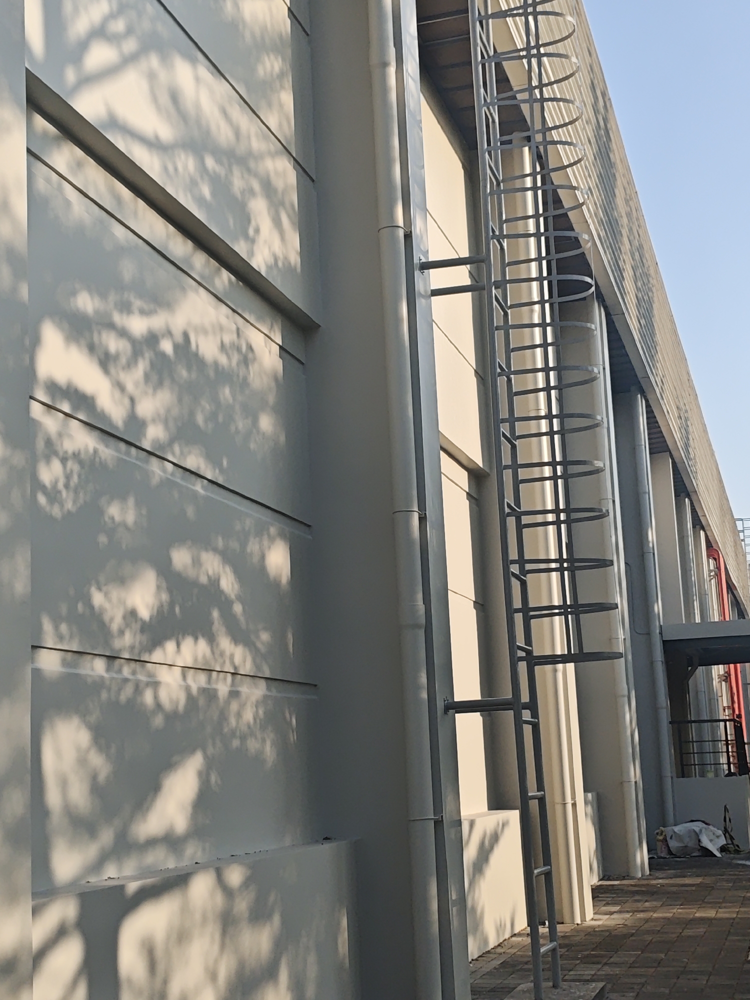 The image shows the exterior of a modern industrial building in Bae, Indonesia. A tall, grey metal ladder with circular safety cages is attached to the side of the building, extending upwards. The building itself has a pale, smooth facade with horizontal lines, casting dramatic shadows from what appears to be tree branches. Along the building's edge, a corrugated metal roof is visible, supported by vertical beams. Several large, grey pipes run vertically down the side of the building. The ground is paved with interlocking brick tiles, and in the background, a covered walkway or bridge structure can be seen. A white tarp covers some objects in the lower right corner of the image. The sky is clear and blue. The overall scene suggests an outdoor industrial or commercial setting.
