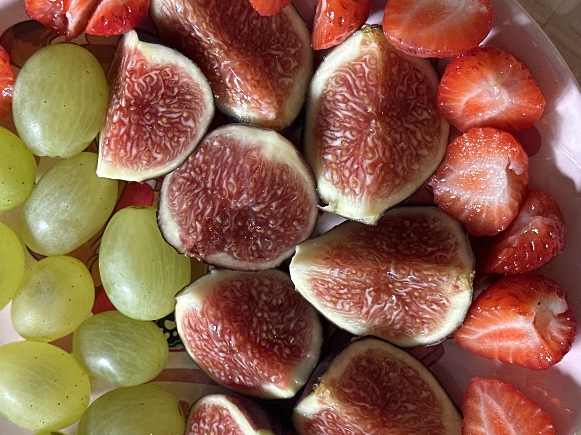 The image displays a close-up, overhead view of a platter filled with sliced fruits. The fruits include several halved green grapes, which are oval in shape and have a slightly translucent, pale green skin. There are also numerous slices of figs, characterized by their vibrant pink and red interiors with visible seeds arranged in a circular pattern. The figs are cut in half, revealing their intricate flesh. Interspersed among the figs are slices of bright red strawberries, each showing its characteristic texture and seeds. The fruits are arranged on a light pink surface, possibly a plate or platter, which has subtle striped patterns in shades of beige and pink. The lighting appears to be natural, creating soft shadows and highlighting the textures of the fruits. The scene is a still life composition of fresh fruit. There is no indication of the city of Wisła, Poland, nor any people or actions.