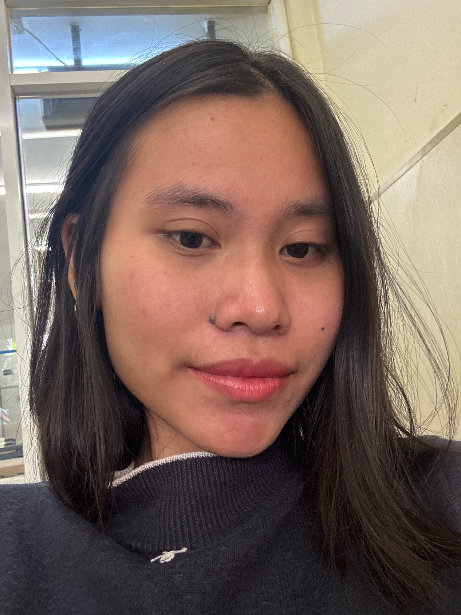 A young woman with dark hair and a dark blue sweater is pictured from the chest up, looking at the camera. She has a small mole on her left cheek and a piercing just below her lower lip. The background appears to be an indoor setting with a tiled wall and a window with horizontal blinds. The lighting suggests it is daytime. There are no discernible elements in the image that would place the location in East Jakarta, Indonesia.