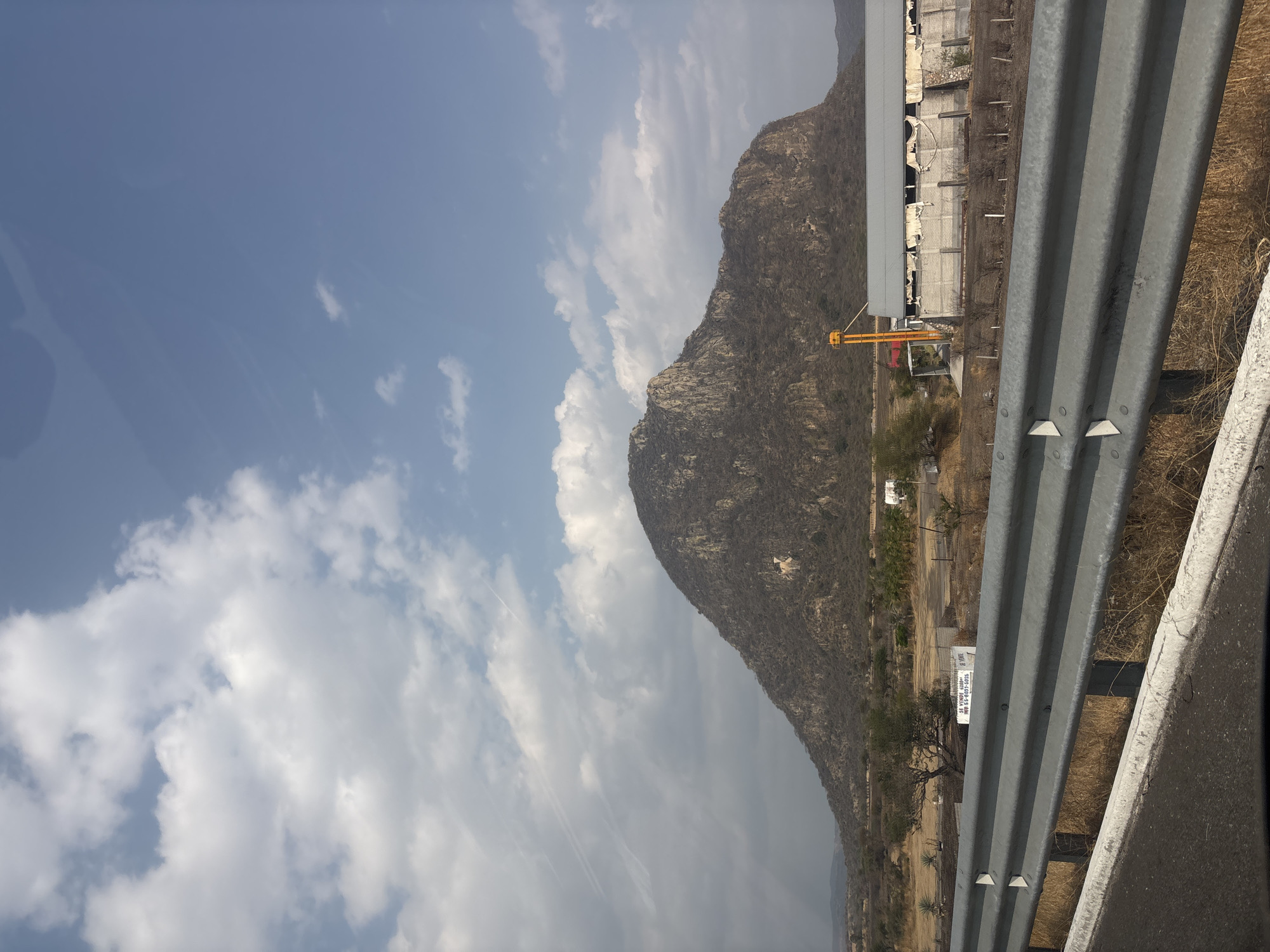 The image captures a view from what appears to be a moving vehicle, focusing on a mountain landscape alongside a highway. The foreground is dominated by a metal guardrail, indicating the roadside.
To the left of the guardrail, a large, rocky mountain ascends, partially obscured by clouds. The sky is visible above and around the mountain, displaying a mix of blue sky and white clouds.
Adjacent to the base of the mountain, along the roadside, there is evidence of human development. A building with signs of disrepair sits close to the mountain's edge. Further down, sparse vegetation and hints of infrastructure suggest the presence of a road.
Based on the location provided, it can be assumed this scene is located in or near Jantetelco, Mexico. However, there are no people visible in the image, and details about specific actions or interactions are absent.