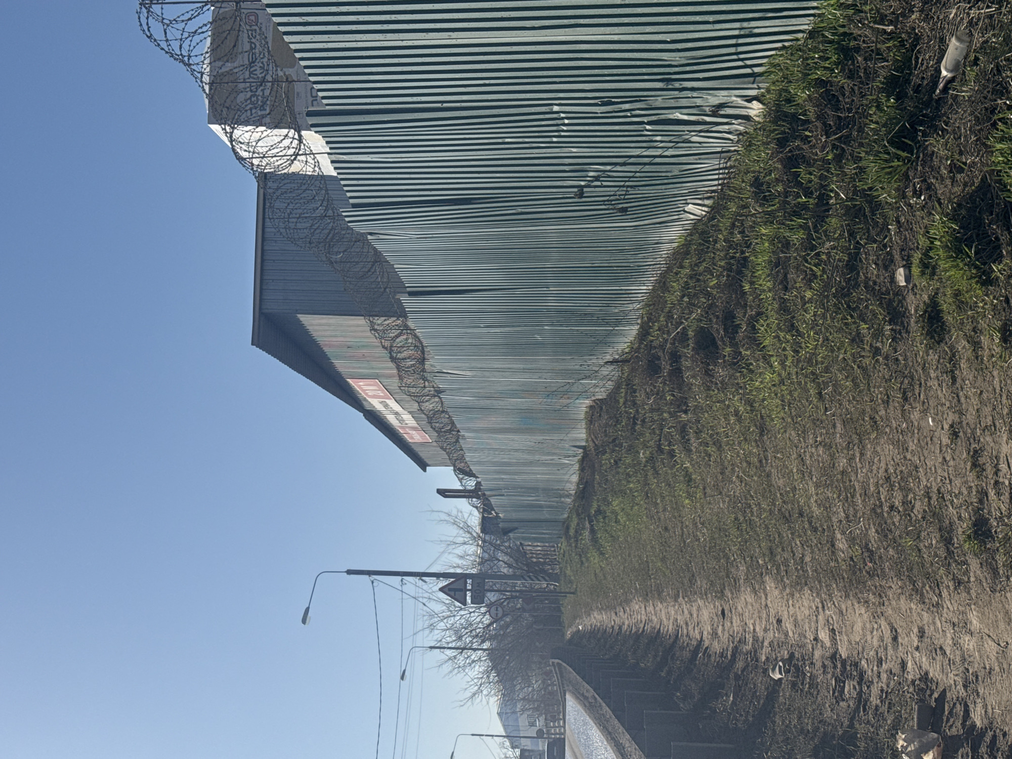 The image depicts a fence topped with barbed wire, running alongside a dirt path overgrown with vegetation. The fence appears to be constructed of corrugated metal, and shows signs of damage. A set of railway tracks are visible in the lower part of the image, suggesting the location may be near a rail line. Bare tree branches and streetlights are present on the opposite side of the tracks. The sky is clear and blue. Based on the prompt information, this is located in the city of Motyakovo, Russia.