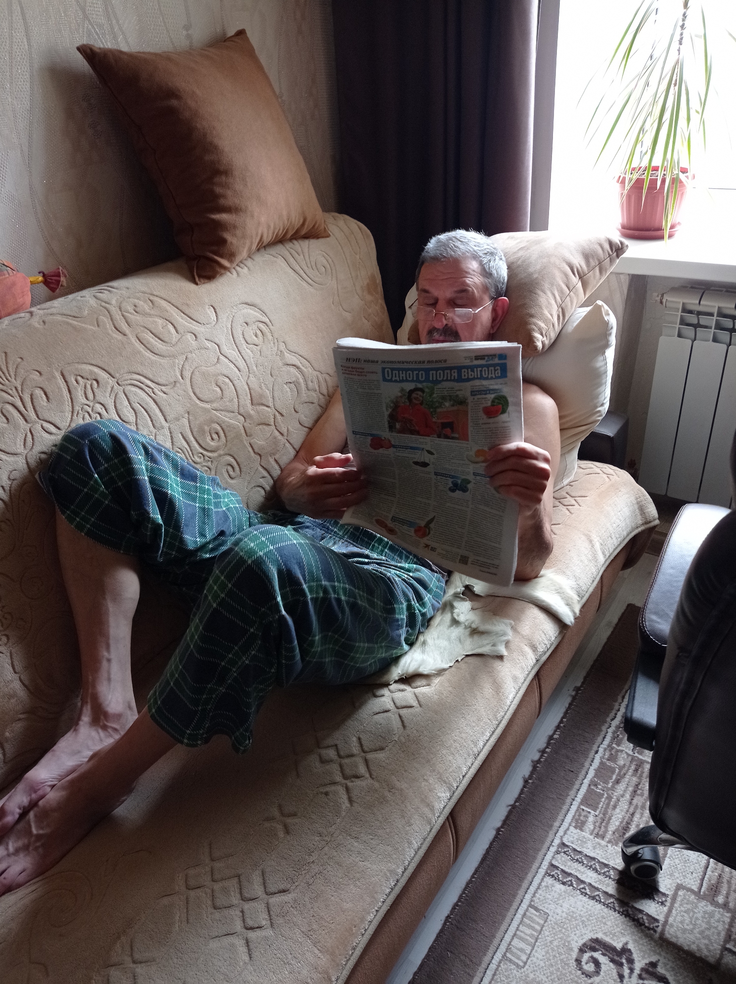 A middle-aged man is reclined on a beige patterned couch in an indoor setting. He is wearing glasses and blue and green plaid pajama pants. He is holding a newspaper with Russian text, the headline reading "ОДНОГО ПОЛЯ ВЫГОДА". The man is barefoot. Behind him are pillows, one brown and one white. Next to the couch is a chair. In the background, a window with a plant in a pot is visible. Based on metadata, the location is Degtyarsk, Russia.