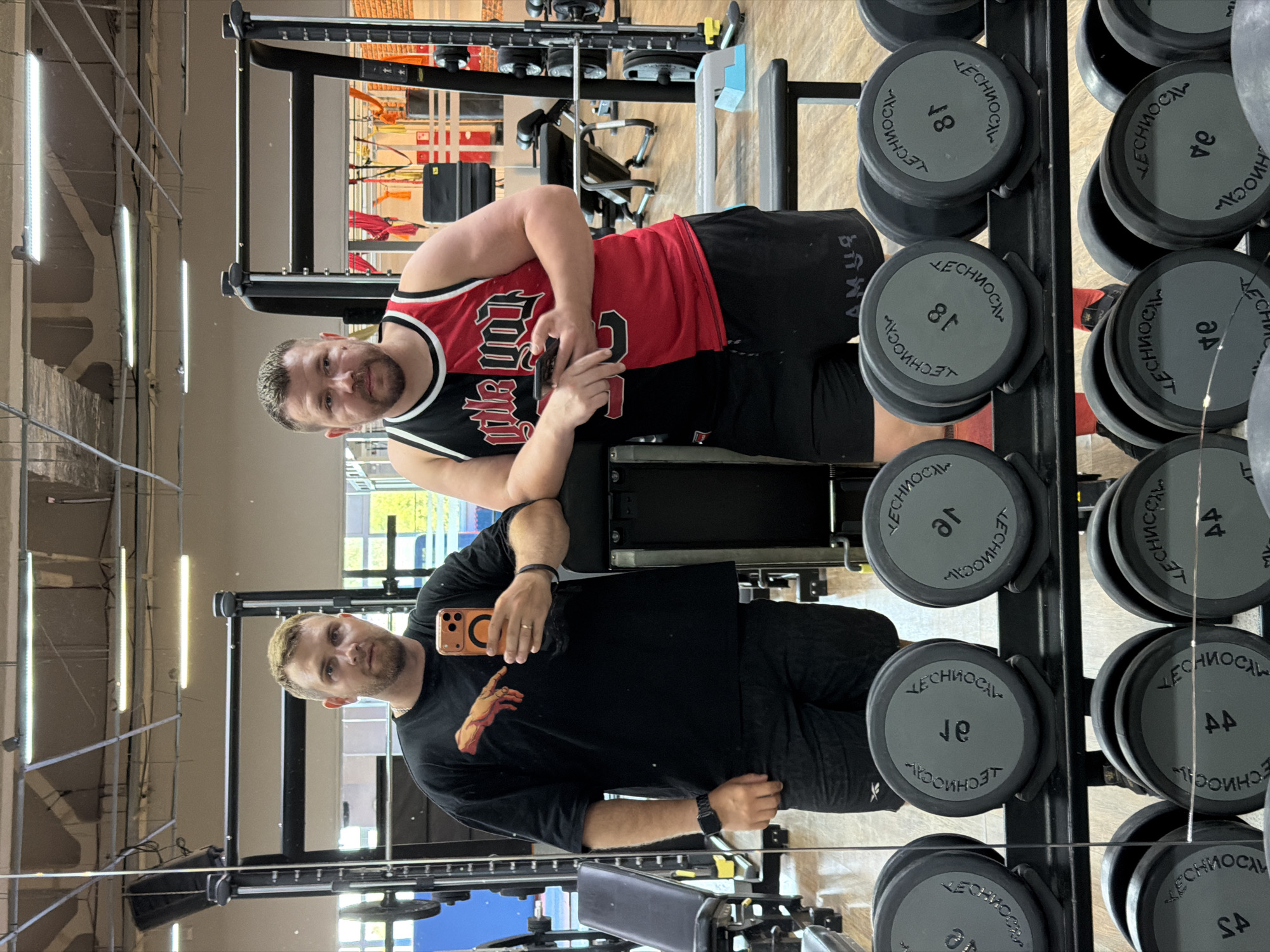 Two men are in a gym, reflected in a large mirror. The man on the left is wearing a black t-shirt with a graphic of a hand reaching out and black shorts. He is holding a smartphone in his right hand, as if taking a selfie. The man on the right is wearing a red and black sleeveless jersey and black shorts. He is holding a smartphone in his left hand. Both men appear to be in good physical condition. The gym is equipped with various exercise machines and weight racks. In the foreground of the reflection, a rack of dumbbells is visible. The lighting in the gym appears to be fluorescent. The scene is captured from a medium shot. No specific geographical markers place this image in Chaikovsky, Russia.