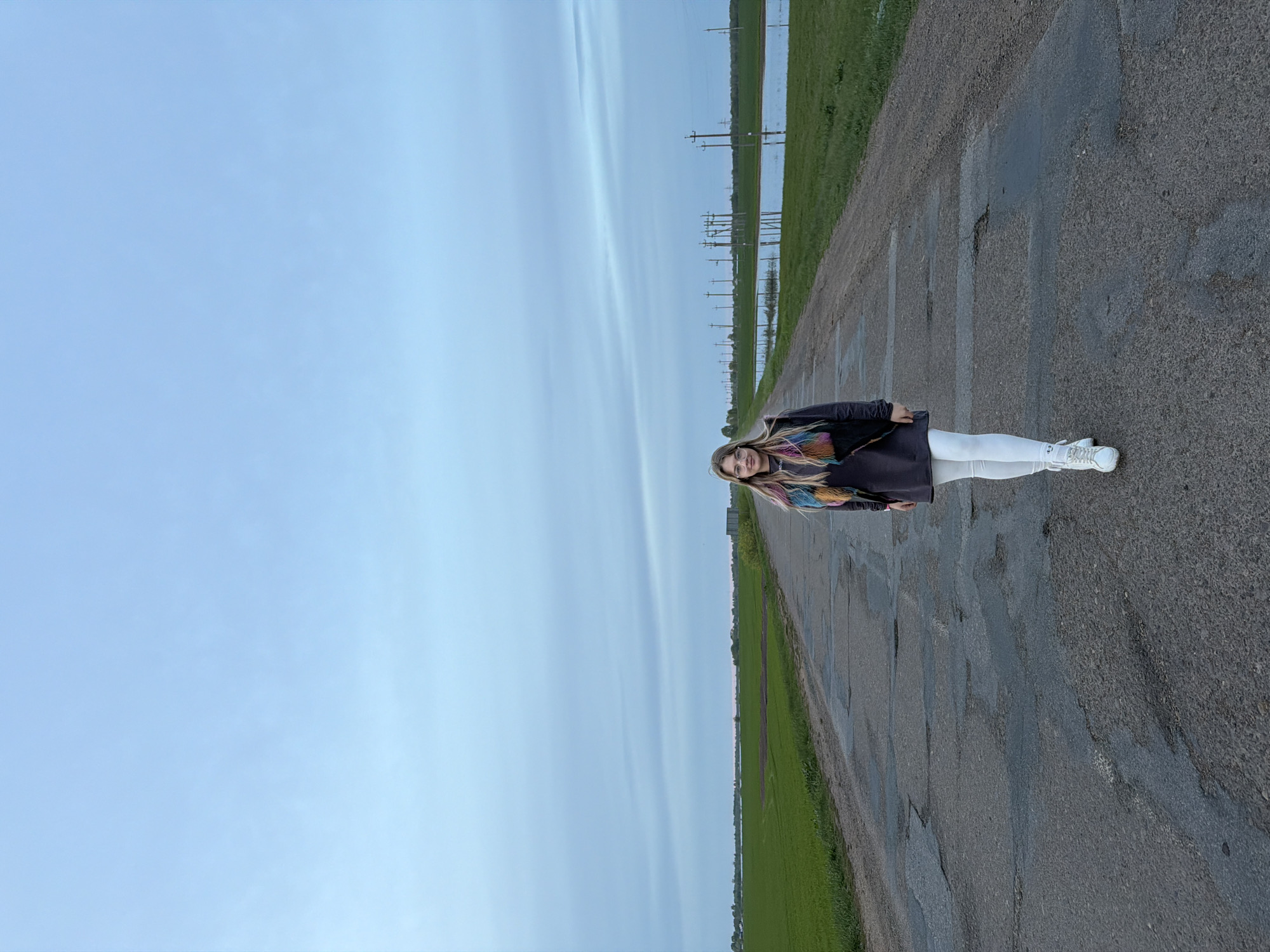 A woman stands on an asphalt road in Azyakul', Russia. The road is adjacent to green fields. Overhead power lines are visible. The sky is overcast. The woman wears white pants, a dark jacket, and sneakers. She has long blonde hair.