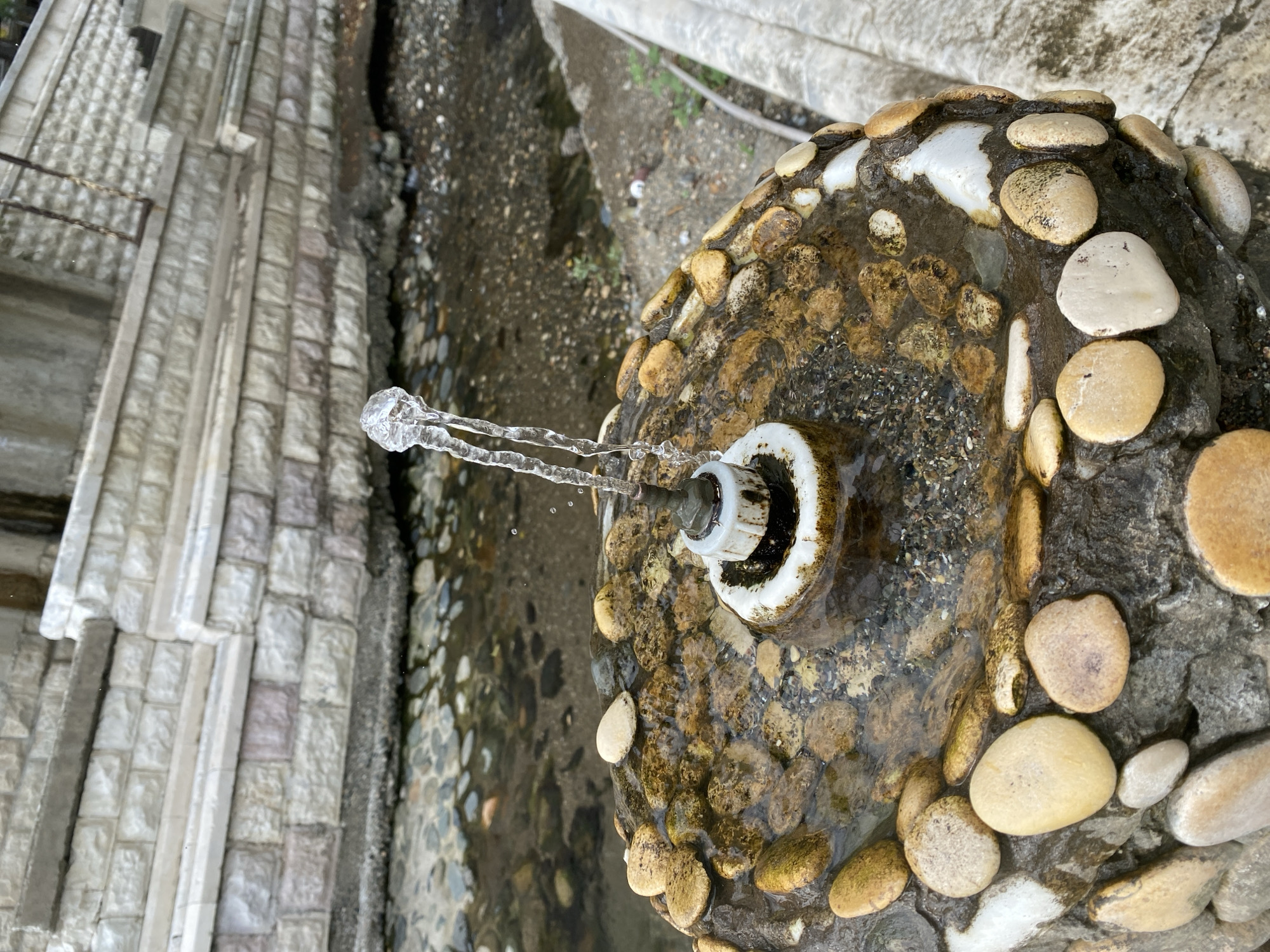 An outdoor close-up shot captures a small, decorative water fountain. The fountain's base is a round, rocky structure covered with smooth, light-colored pebbles and some darker, wet stones. A white ceramic fixture sits in the center of the fountain, from which a thin stream of water emerges, bending and twisting as it falls back into the basin. The water is clear and shows its movement with a few small droplets in the air. Behind the fountain, a shallow stream with a gravelly bottom is visible, and beyond that, a textured stone wall with rectangular blocks forms a raised embankment. The overall scene suggests a park or a public space with natural elements. The location is Sokhumi, Abkhazia.
