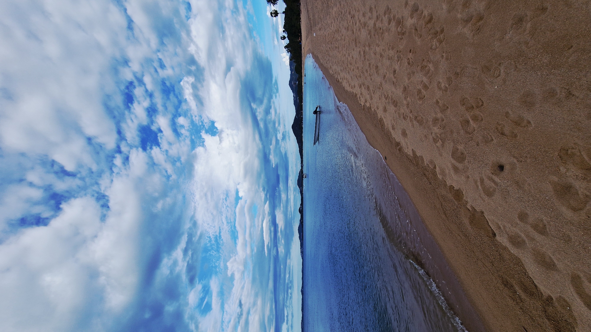 The image shows a beach scene in Baan Tai, Thailand. The foreground is a sandy beach with numerous footprints. To the left, the ocean meets the shore with gentle waves. In the water, a small white boat with an outboard motor is anchored. In the distance, hills and lush green vegetation line the coast, with a few palm trees visible on the right side of the image. The sky above is filled with dramatic clouds against a blue backdrop. The overall atmosphere is serene and picturesque.