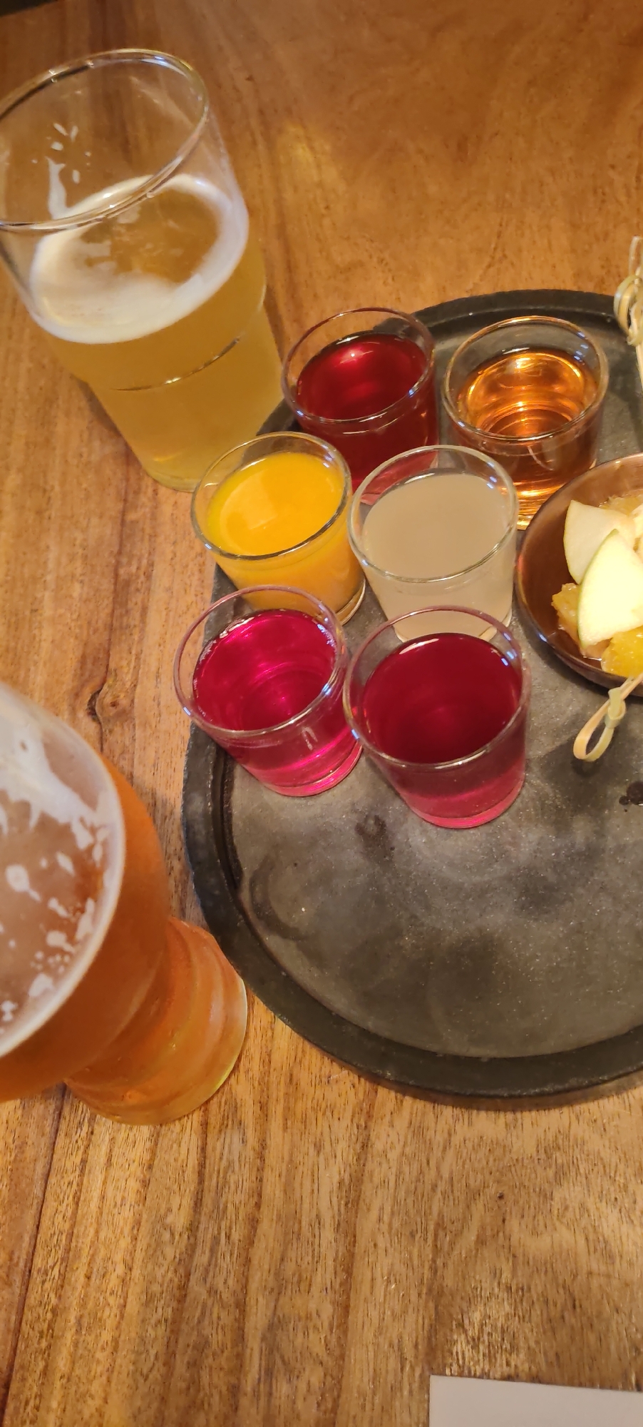 A close-up, overhead shot captures a sampling of beverages and some fruit on a wooden surface. A tall glass of pale yellow beer with a frothy head stands to the left, partially obscuring a dark, circular tray. On the tray, arranged in a semi-circle, are six small shot glasses filled with various colored liquids: two dark red, one orange, one pale yellow, and two more dark red. A small, shallow dish to the right holds slices of green apple and orange segments, speared by a twisted twig. The lighting is warm and the focus is sharp on the beverages. The image does not contain any information about the city of Kaluga, Russia.