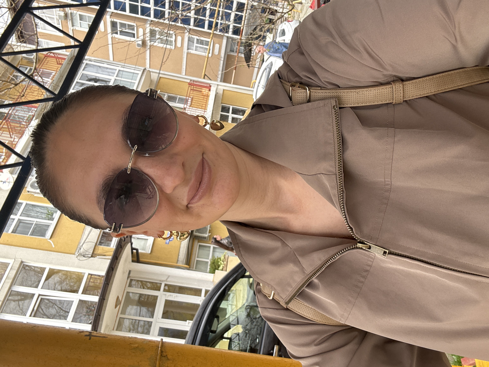 A woman takes a selfie outside in the city of Sochi, Russia. The woman is wearing sunglasses and a light brown jacket. Behind the woman is a multi-story building and a tree with bare branches. Part of a black vehicle is also visible. The weather appears to be bright and sunny.
