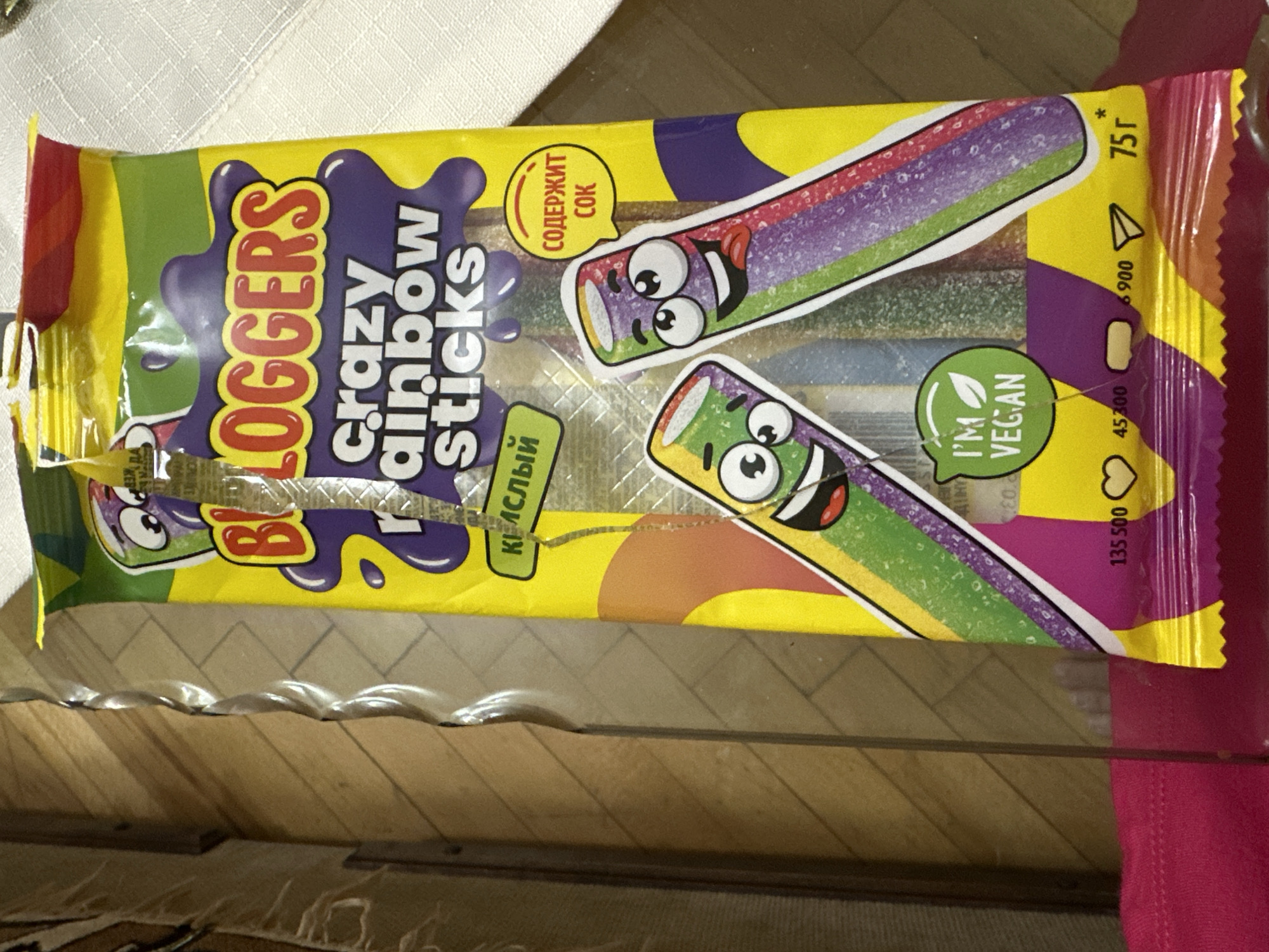 The image is a close-up, eye-level shot of a brightly colored package of "Bloggers Crazy Rainbow Sticks" candy. The package is predominantly yellow with purple, green, and red accents. It features cartoon-like illustrations of two candy sticks with faces. One candy stick is green and purple, with wide eyes and a happy, slightly protruding tongue. The other is green, purple, and red, also with a smiling face. Text on the package includes "Bloggers," "crazy rainbow sticks," "Кислый" (meaning sour in Russian), "СОДЕРЖИТ СОК" (meaning contains juice in Russian), and "I'M VEGAN" with a vegan symbol. There are also numerical markings and icons at the bottom of the package, including a heart symbol, a paper airplane, and "75г" (meaning 75 grams). The packaging appears slightly wrinkled and has a transparent plastic overlay. The candy sticks themselves are visible through the packaging, showing distinct rainbow layers of color. The background is blurred, showing a wooden surface with diagonal lines, possibly a floor or table, and a white textured surface, likely a tablecloth. The lighting is diffuse, creating soft shadows. There are no people or discernable landmarks to indicate a specific location in Yerevan, Armenia. The image is solely focused on the product packaging.