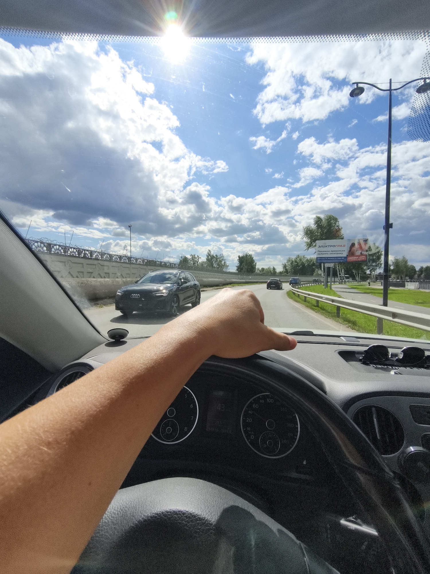 The image captures a first-person perspective from inside a car, looking forward through the windshield. A person's arm and hand are visible, resting on the steering wheel, indicating they are driving. The car is on a paved road, with other vehicles in front and to the side.
The scene is set outdoors on a bright, sunny day with scattered white clouds in a blue sky. The sun is high in the sky, suggesting it is daytime, likely midday. The presence of green grass and trees lining the road indicates a summer or late spring setting.
Notable details include a large billboard on the right side of the road with Russian text and images, advertising "ЭЛЕКТРОГРАД" (Elektrograd) and offering a "СКИДКА 10%" (10% discount). A streetlamp is also visible along the roadside. The dashboard of the car is partially visible, showing a speedometer and other indicators. The road appears to be in an urban or suburban area, given the presence of infrastructure like the billboard and lampposts, as well as the style of the wall and fencing on the left side of the road.
The overall mood conveyed is one of a typical driving experience on a pleasant day, with the focus on the journey and the surrounding environment. There are no explicit indicators of strong emotions, but the bright sunlight and clear sky suggest a generally positive or neutral atmosphere.