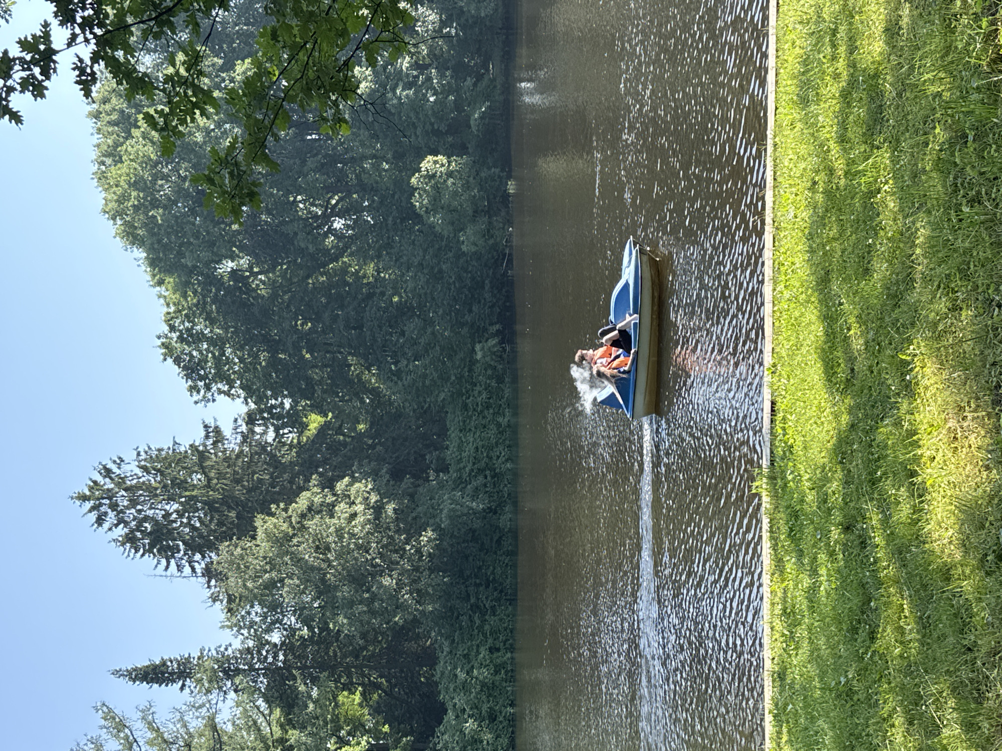 A bright, sunny day is depicted in this outdoor scene. The photograph is oriented vertically, showcasing a tranquil body of water with a lush green grassy bank to the right and dense trees lining the left side. The water reflects the sunlight, creating a dappled effect.
In the middle of the water, a small blue pedal boat is moving, leaving a wake behind it. Two individuals are seated in the boat, both wearing orange life vests, suggesting they are engaged in a recreational activity. One person appears to be pedaling, while the other is looking forward. A small amount of mist or spray is visible around the boat, possibly from the pedaling action or movement through the water.
The trees on the left are vibrant green and appear to be in full leaf, indicating a warmer season, likely summer. The sky above is a clear, pale blue. The grass on the right is neatly trimmed and illuminated by the sunlight, with some small white flowers visible. The overall atmosphere of the image is peaceful and serene, conveying a sense of leisure and enjoyment of nature. The location is Saint Petersburg, Russia. There is no visible text in the image.