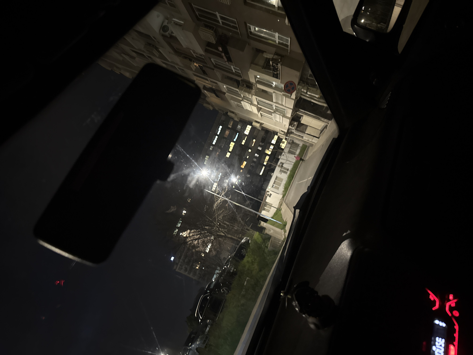 The image shows a nighttime scene from inside a car in Burgas, Bulgaria. The car's interior is visible in the foreground, with parts of the dashboard and rearview mirror discernible. Through the car window, a view of an urban landscape is visible. A tall apartment building with lit windows dominates the scene. Streetlights illuminate the street and surrounding greenery. Several parked cars are visible along the road. A traffic sign is mounted on the building, which could indicate the approximate location. The building is likely located in a residential area of Burgas.
