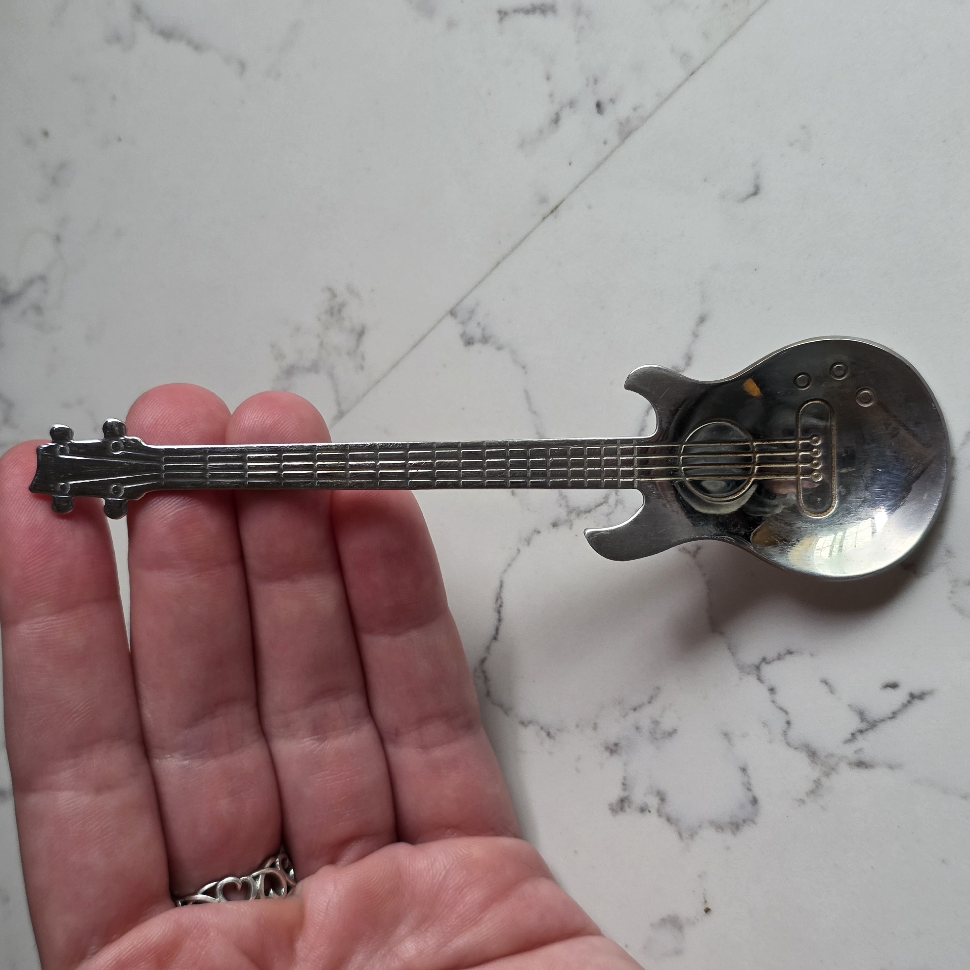 In Beaverton, Oregon, a hand with a decorative ring is holding a silver spoon shaped like a guitar. The spoon has a detailed design, including a headstock with tuning pegs, a fretted neck, a body with a soundhole and strings, and control knobs. The background is a white marble surface.