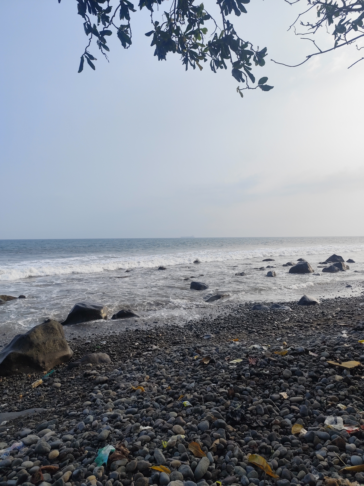 The image depicts a rocky beach in Teba, Indonesia. The foreground is covered in dark grey pebbles and stones, with scattered debris including plastic bags and fallen leaves. Several larger, dark rocks are interspersed among the pebbles, with some partially submerged in the shallow water. The ocean stretches to the horizon, with gentle waves breaking on the shore, creating white foam. The sky is overcast with a light haze, and the branches of a tree with dark green leaves hang into the upper portion of the frame. The overall scene is that of a natural, albeit somewhat littered, coastal environment.