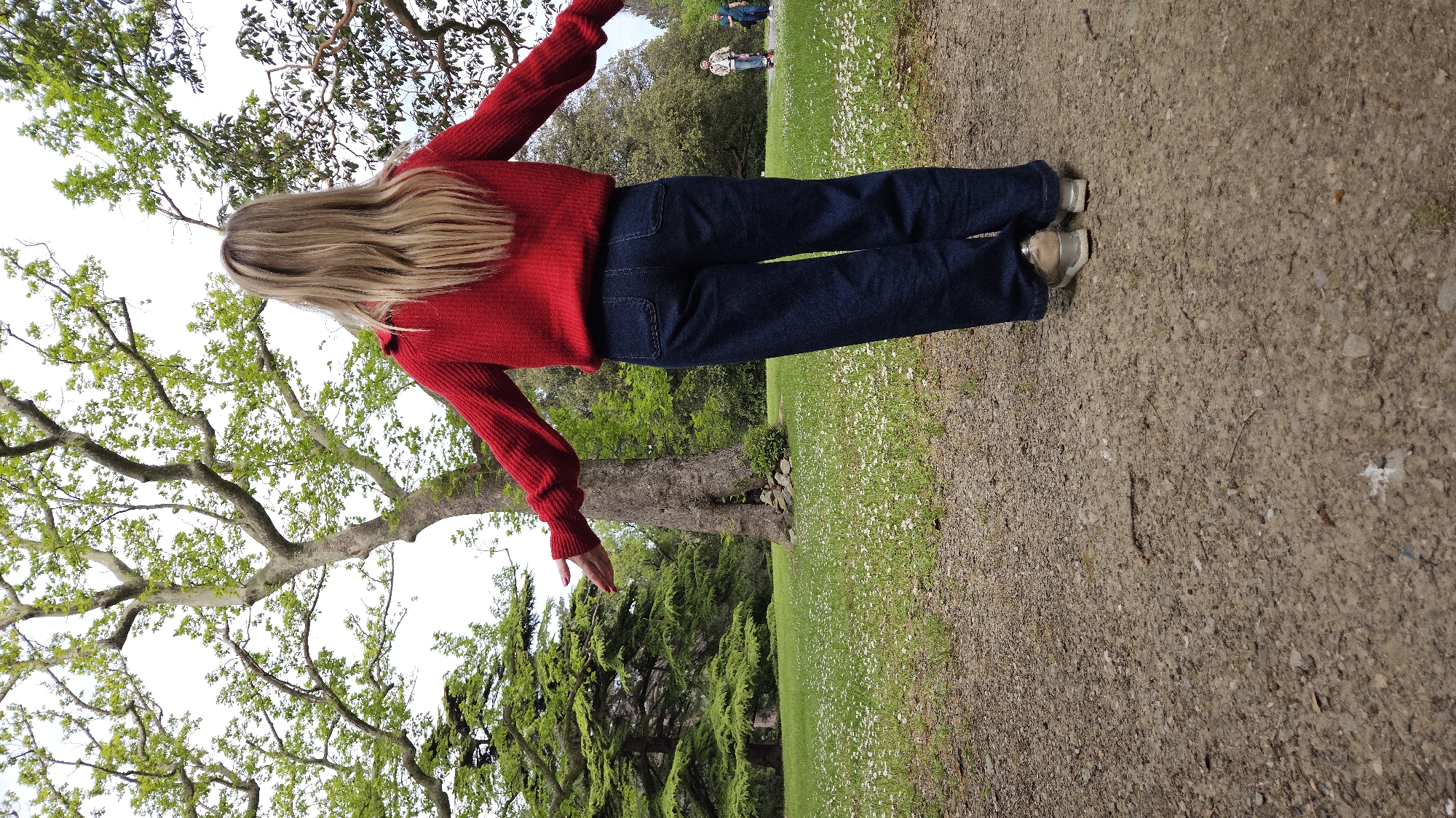 A woman stands on a dirt path with grass and small white flowers on either side in Unknown, Unknown. She wears a red sweater, blue jeans, and light-colored shoes. Her blonde hair falls down her back. Her arms are outstretched to either side. Trees are visible on both sides of the path. In the background, two people stand on the grass.