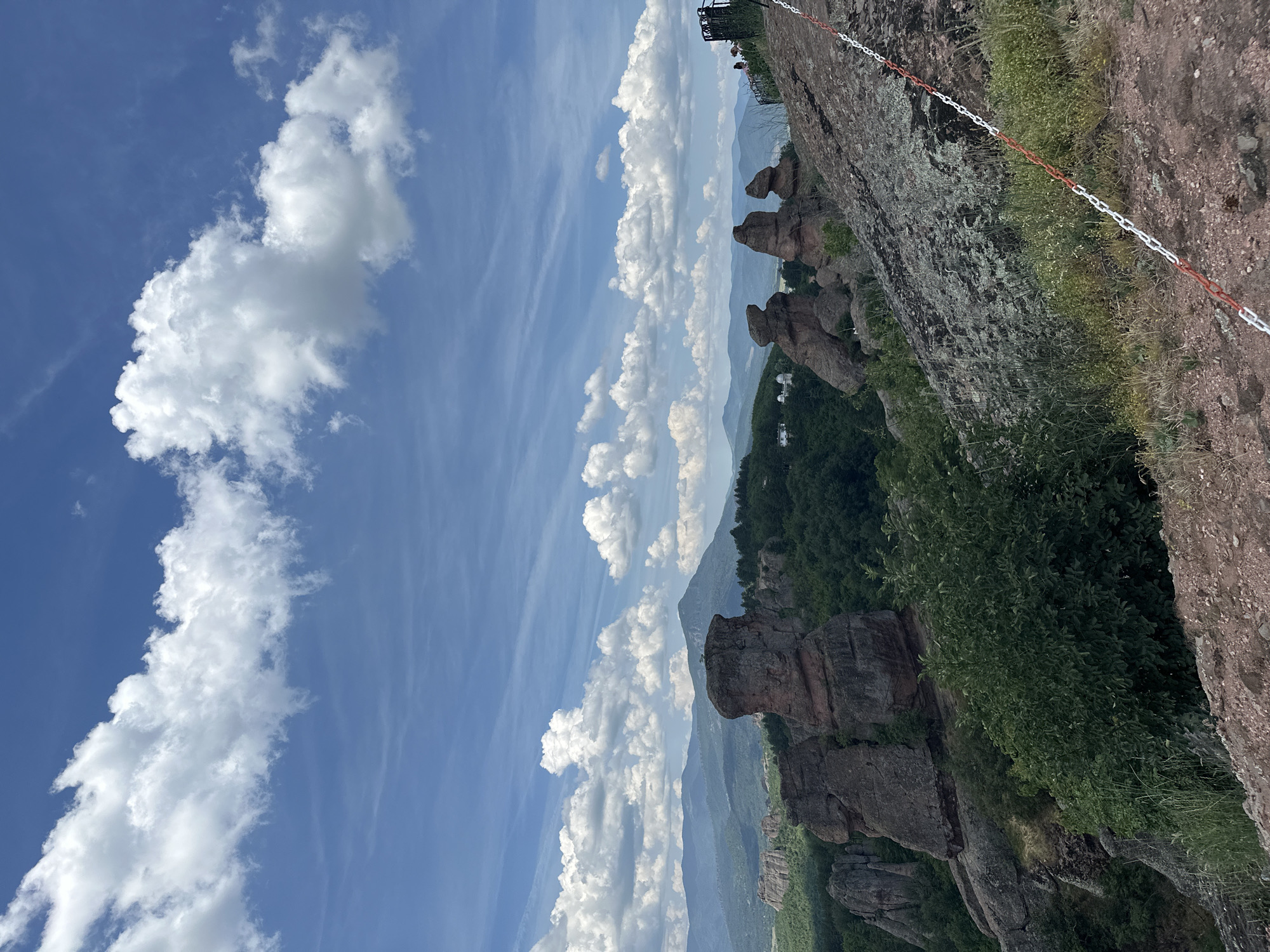 The image depicts a natural landscape dominated by striking rock formations in the city of Belogradchik, Bulgaria. In the foreground, a rugged, rocky path leads up and to the right, marked by a red and white chain barrier. The path is edged with dry grass and sparse vegetation. The mid-ground showcases several prominent, weathered sandstone rock pillars, some appearing solitary and others clustered, rising from a lush, green forested area. In the distance, rolling hills and mountains form a layered backdrop under a bright blue sky dotted with white, fluffy clouds. A few structures, possibly buildings or domes, are visible nestled among the trees on the left side of the frame. On the far right, a railing with a few silhouetted figures can be seen, suggesting a viewing point. The overall scene conveys a sense of natural grandeur and geological uniqueness characteristic of the Belogradchik Rocks.